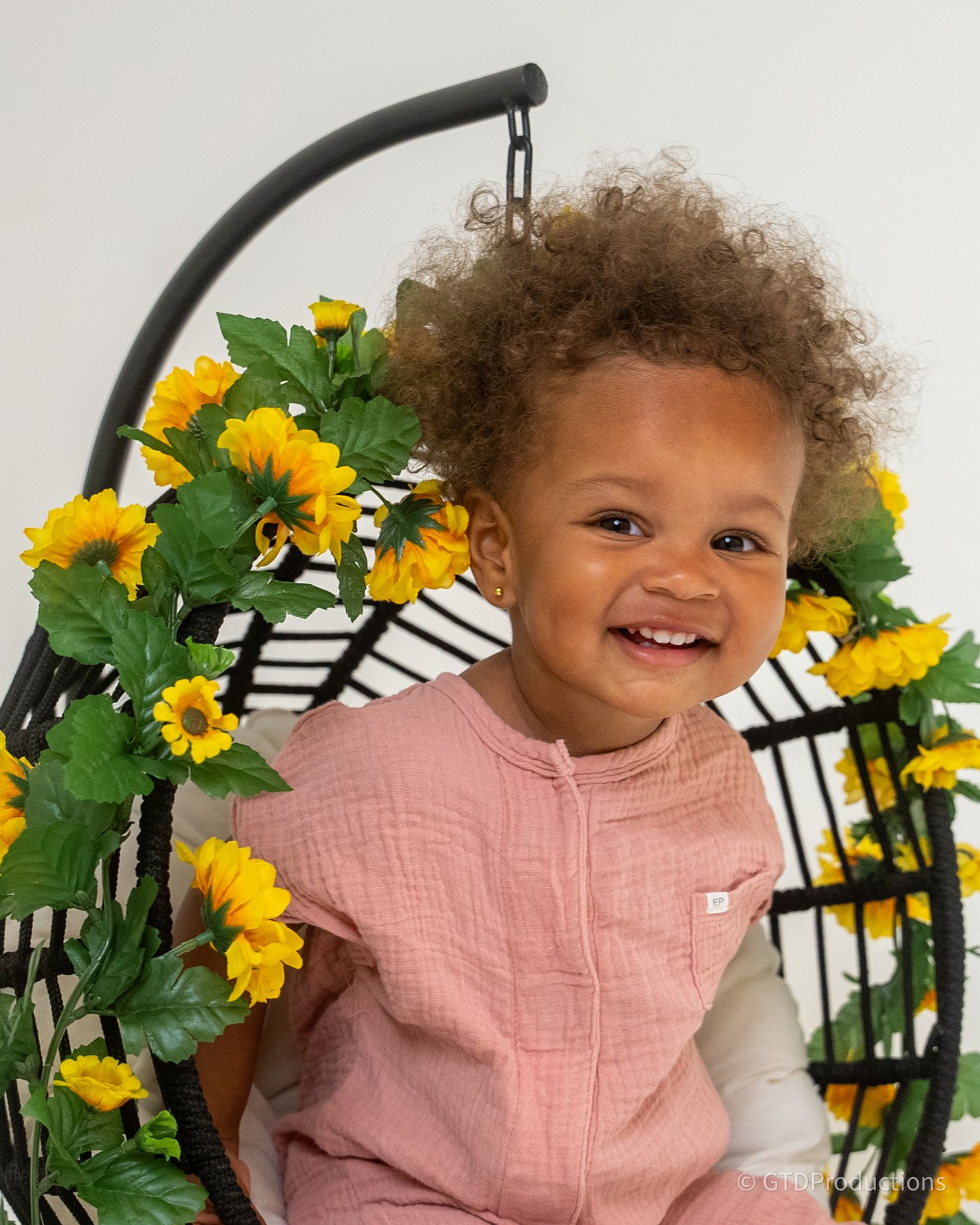 A joyful toddler sitting in a chair decorated with yellow sunflowers against a white background.
