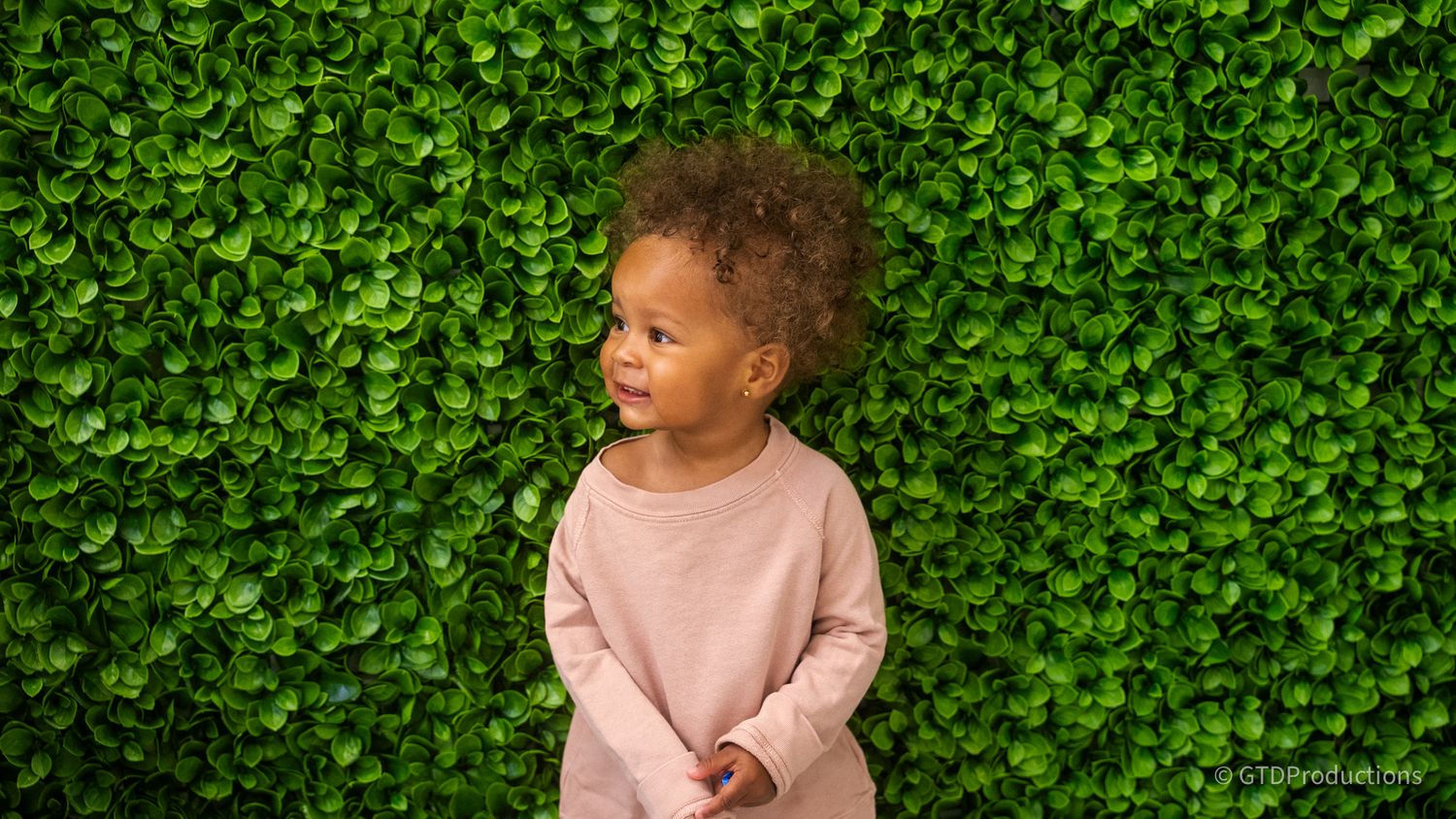A child in pink clothing photographed against a lush green hedge background.