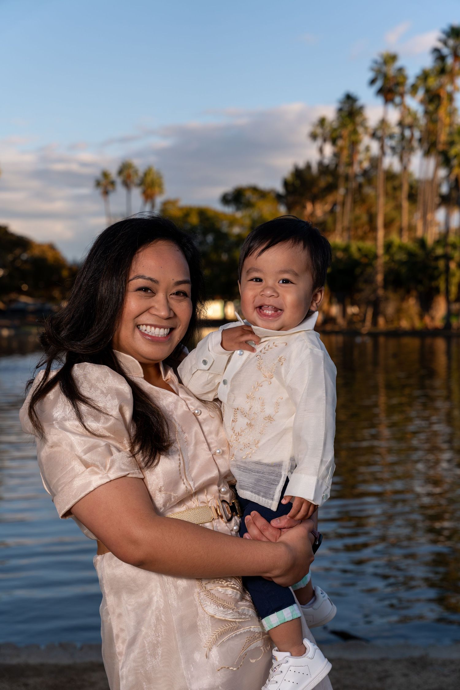 Mother and child in white outfits share joyful moment by waterfront at sunset.
