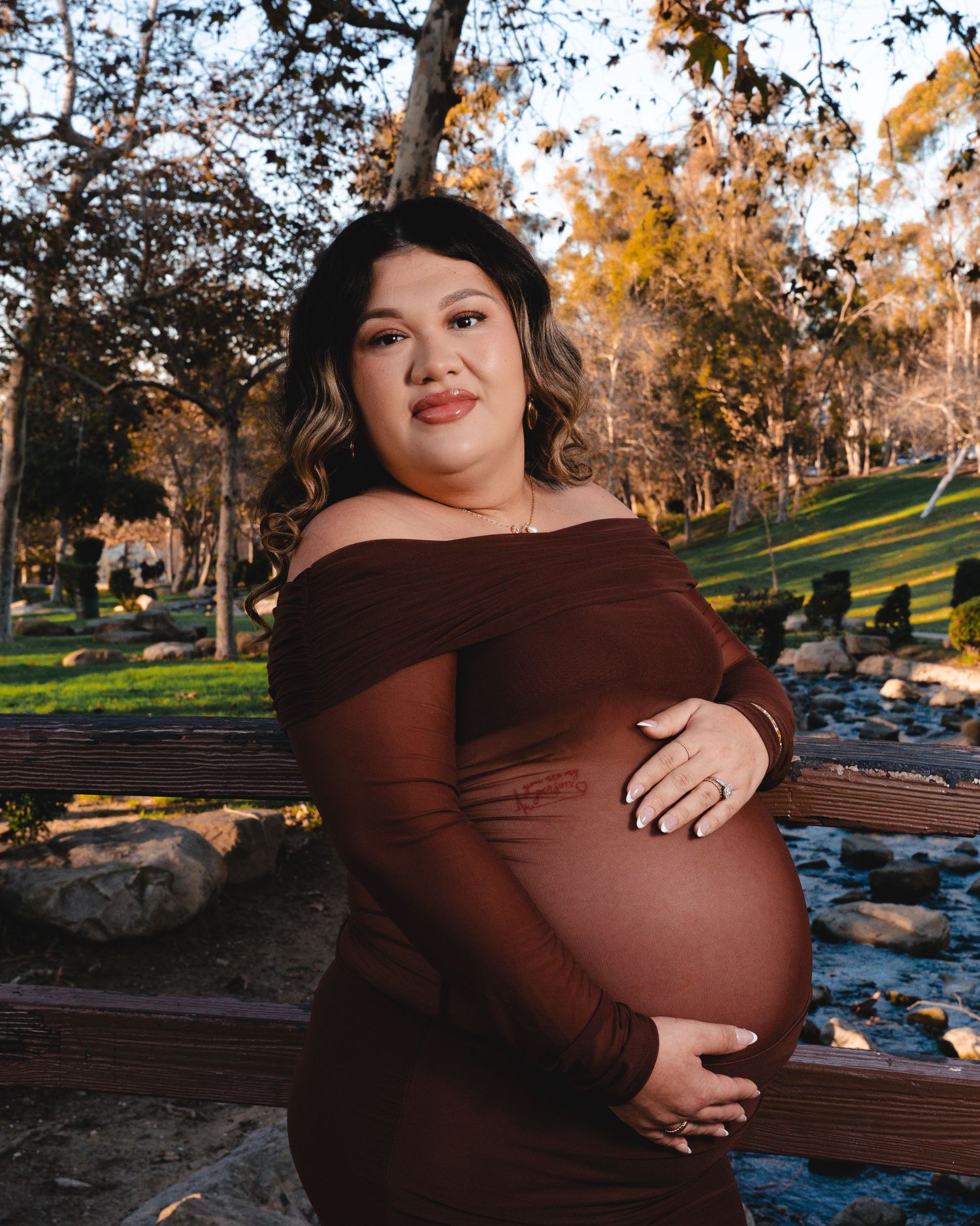 A maternity photoshoot in a park during golden hour with a woman in a brown dress sitting on a bench.