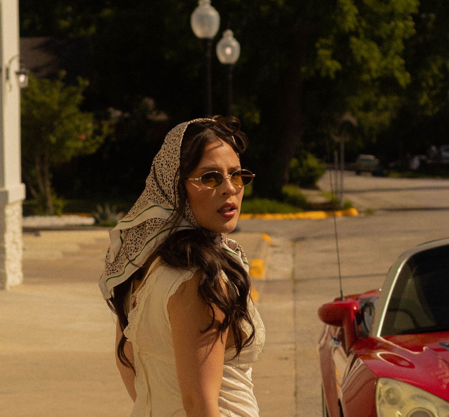 Woman in white dress and patterned headscarf with sunglasses standing near a red car on a sunny day.