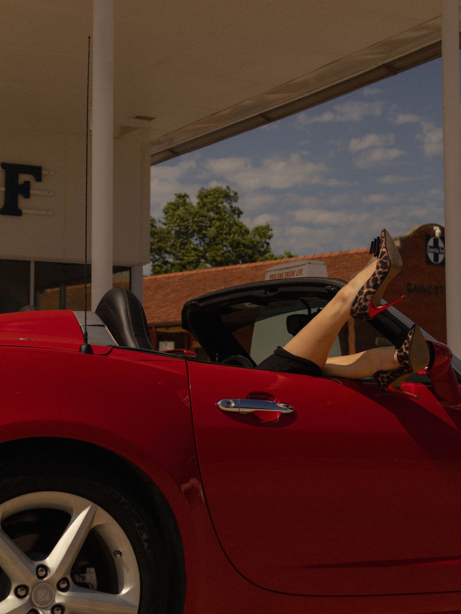 Red convertible sports car parked at a Gulf gas station with a woman's legs resting on the door.
