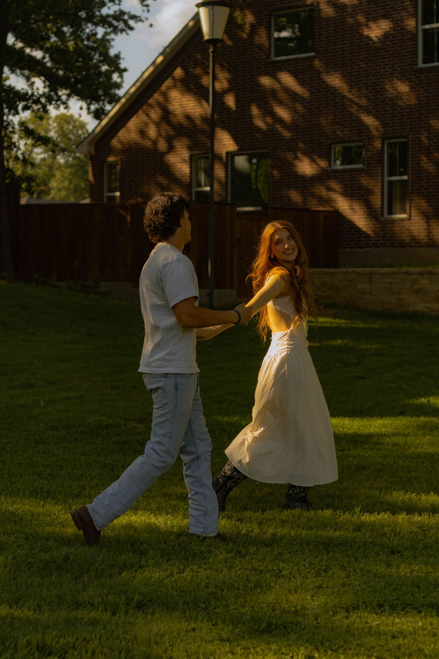 Couple holding hands walking on green grass at golden hour near brick building.