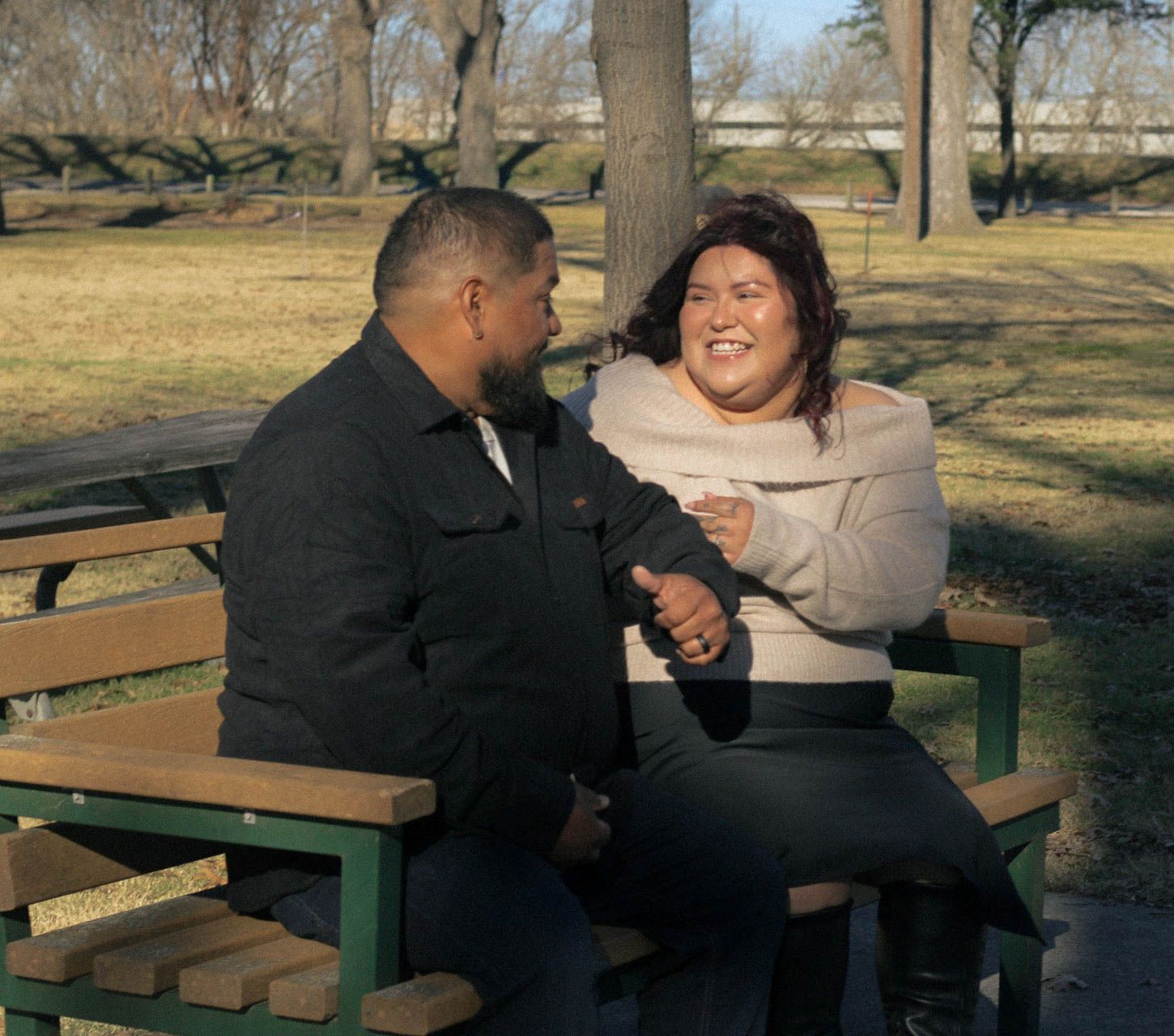 Two people share a lighthearted moment while sitting together at an outdoor bench in a park setting.