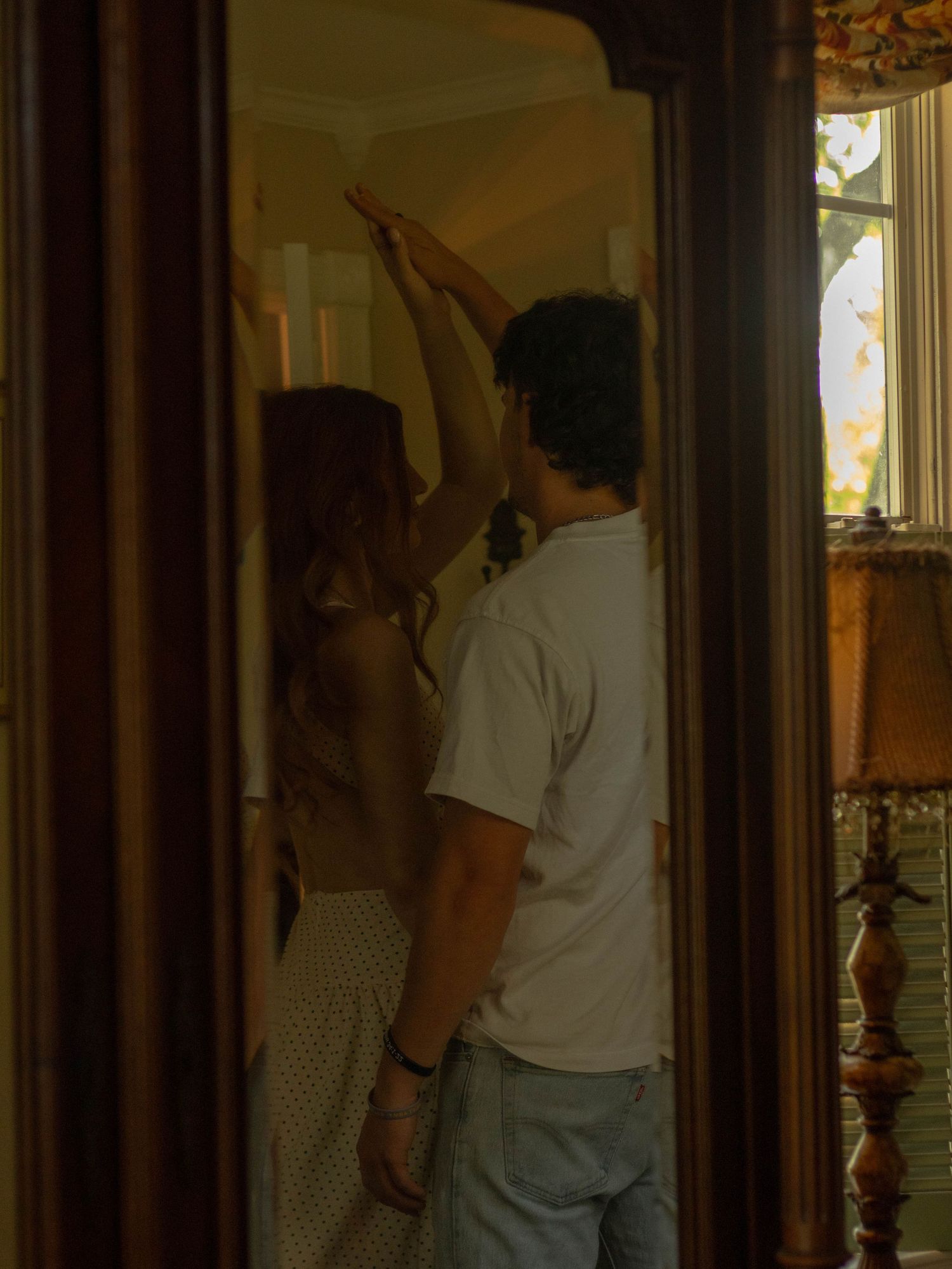 Couple reflected in tall vintage mirror reaching upward together inside a warmly lit historic home.