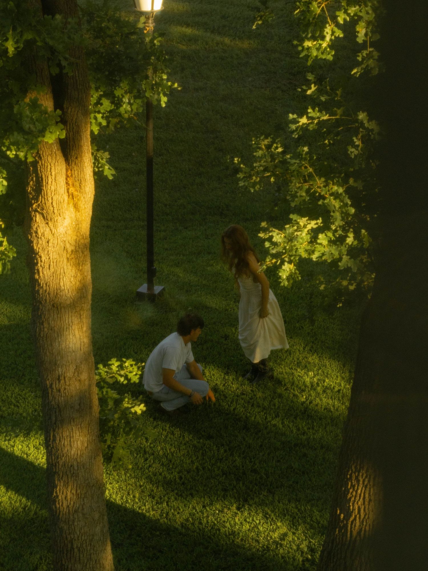 A man crouching and a woman in a white dress standing on lush green grass among trees at dusk.