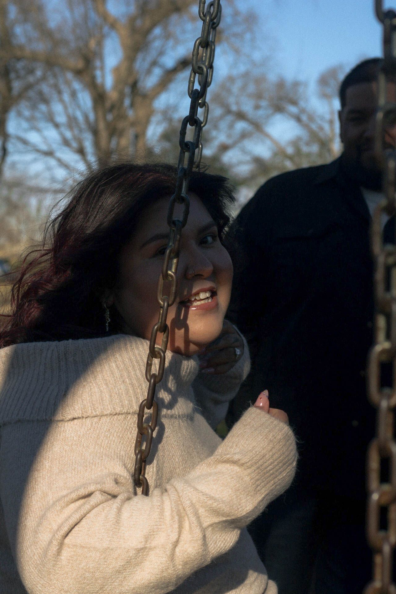 Person smiling while swinging in a park with bare trees in the background.