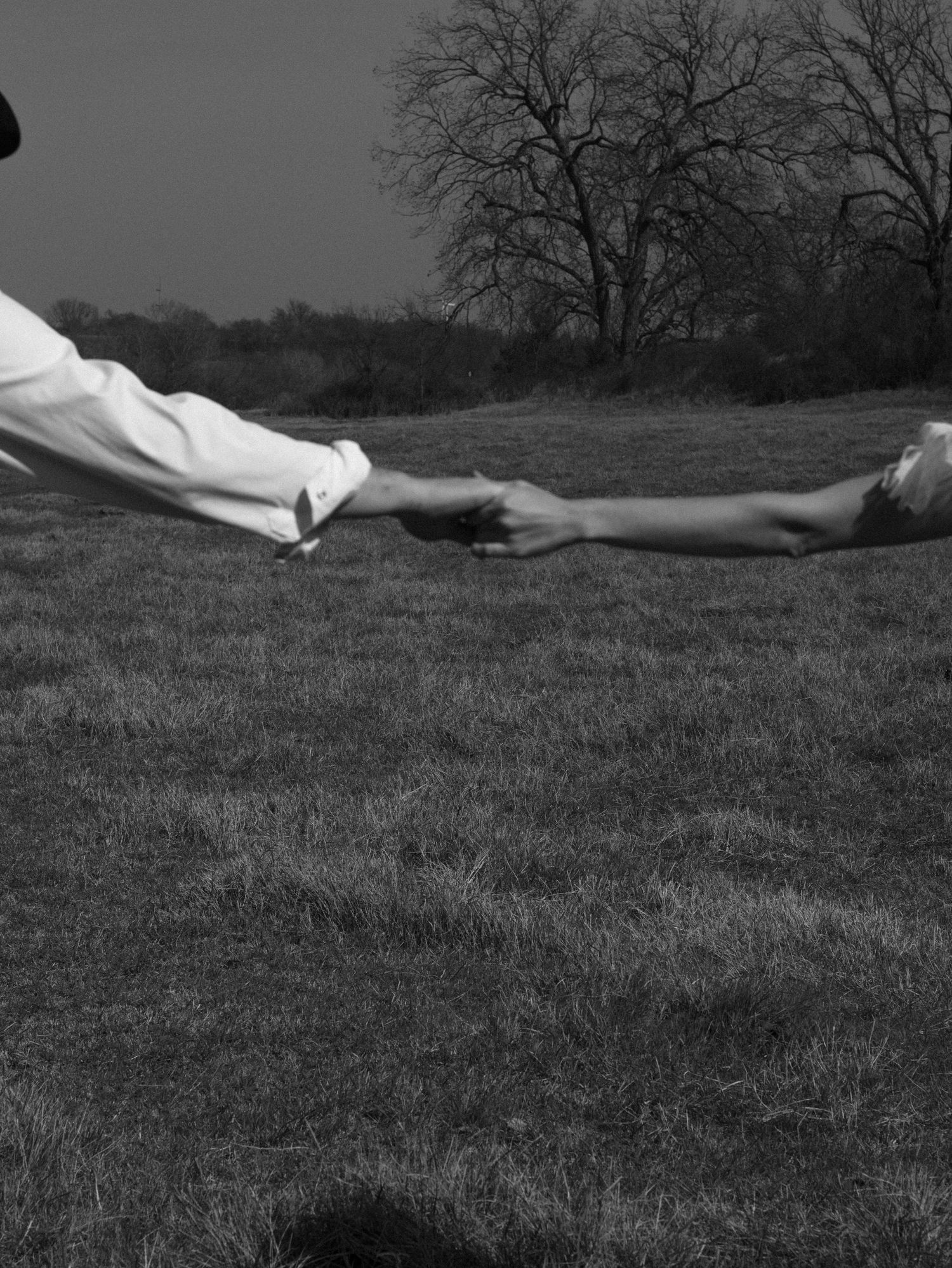 Black and white photo of two people reaching toward each other across a grassy field with bare trees in the background.
