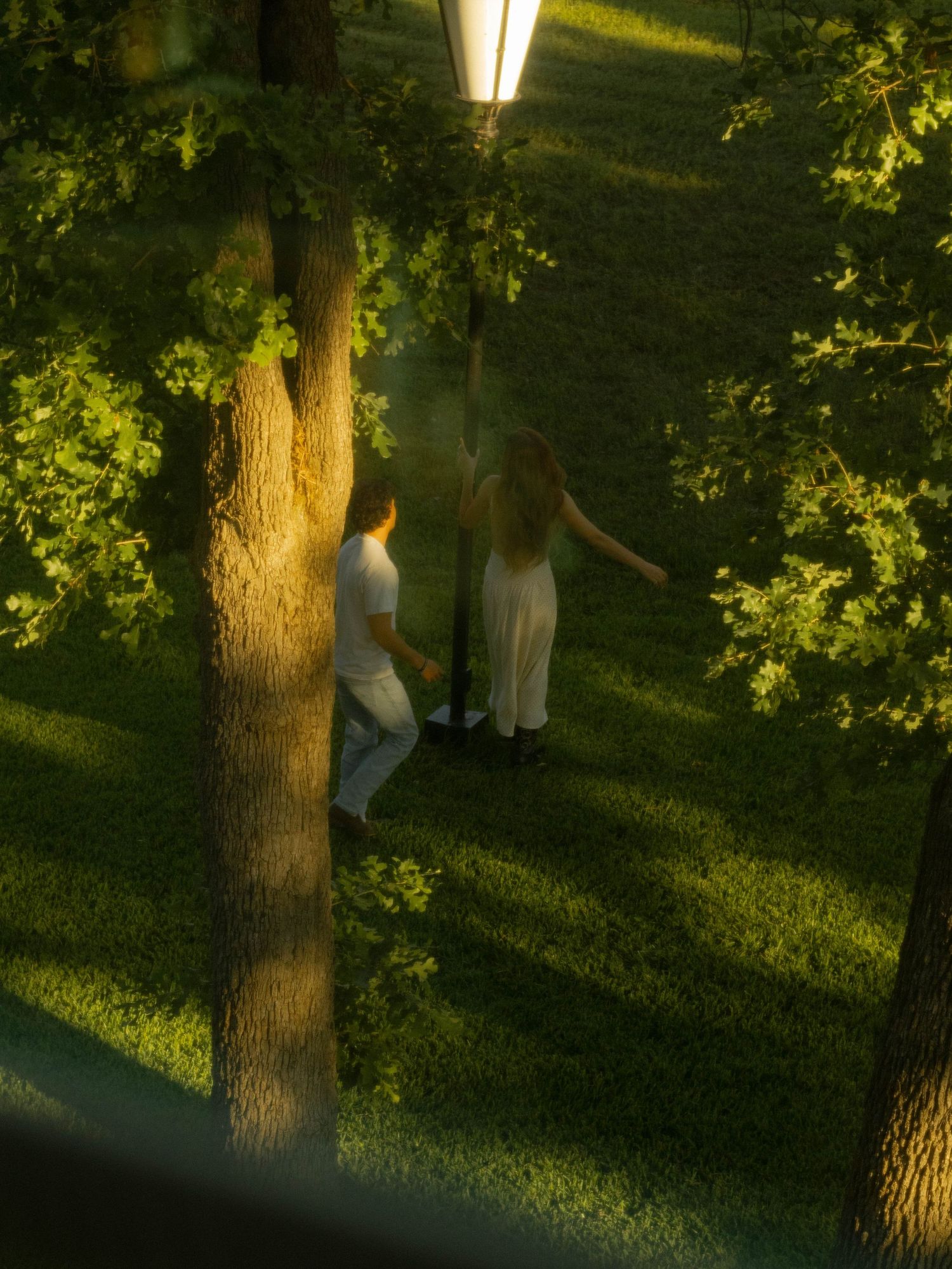 Two people in white clothing walk through a lush green park at dusk, illuminated by a vintage street lamp.