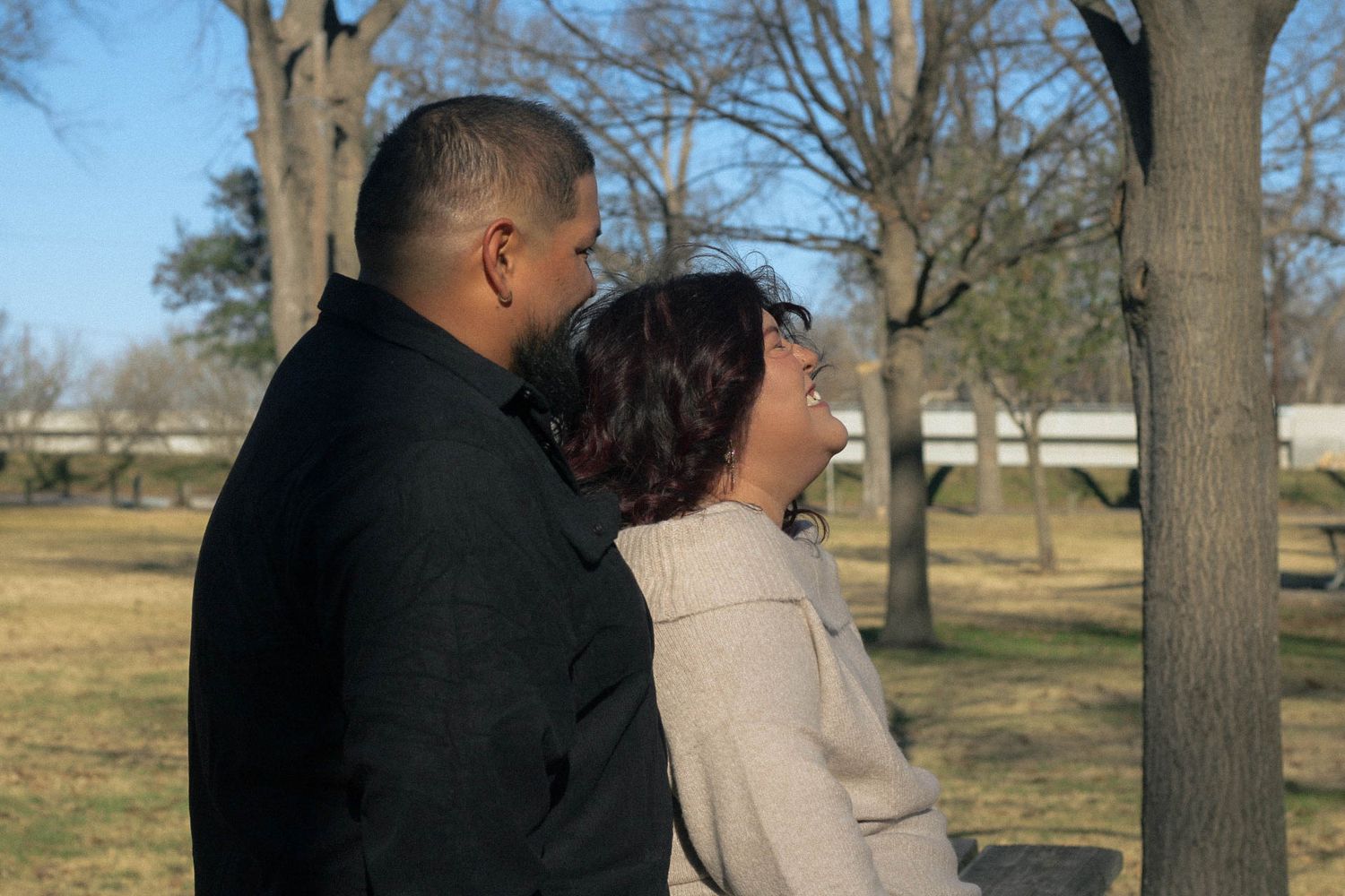 A couple in contrasting outfits poses together against a backdrop of bare trees.