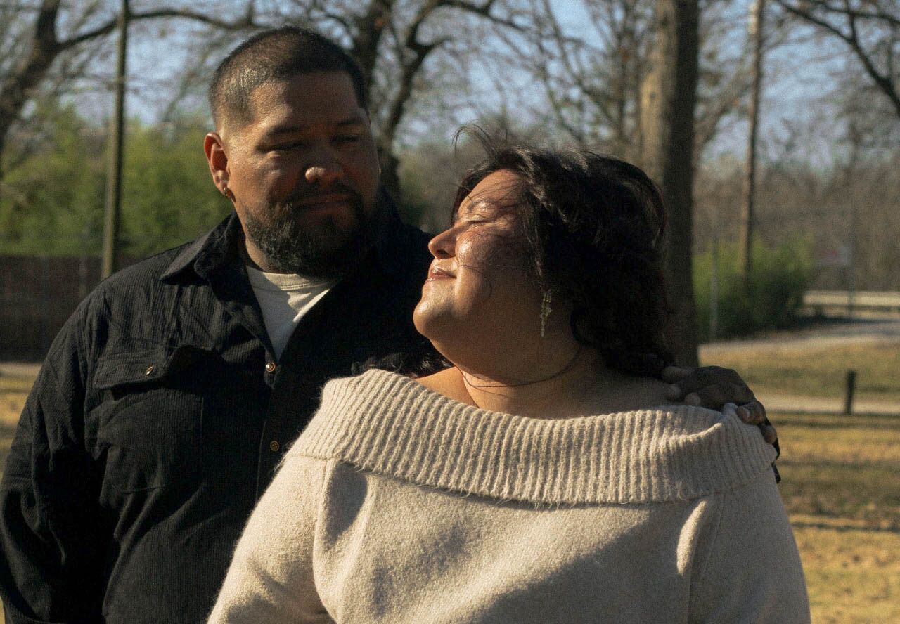 Two people pose together in casual attire during a sunny outdoor photoshoot.