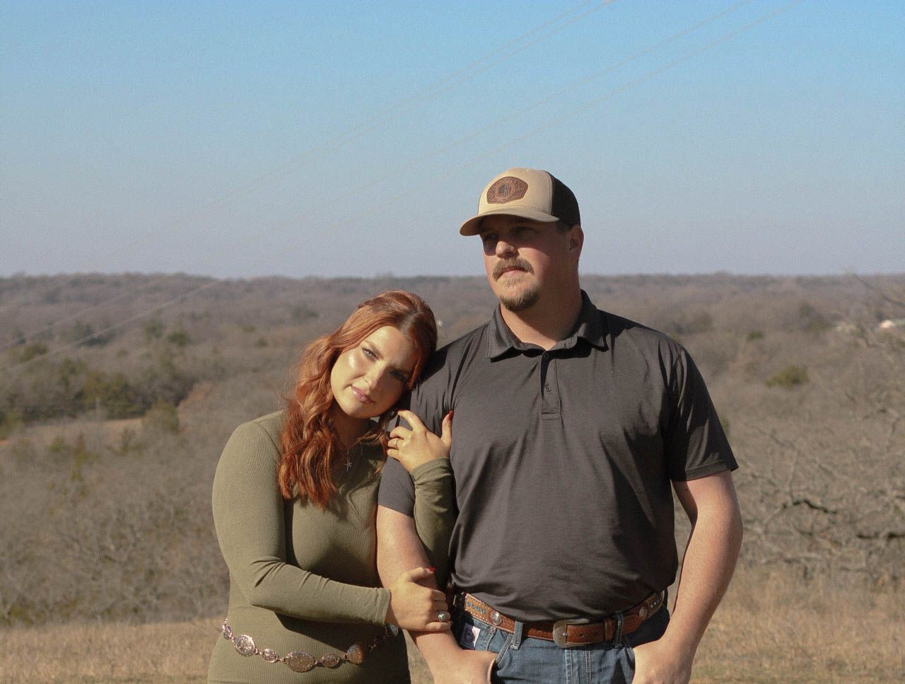 A couple stands together in an arid landscape with hills in the background.