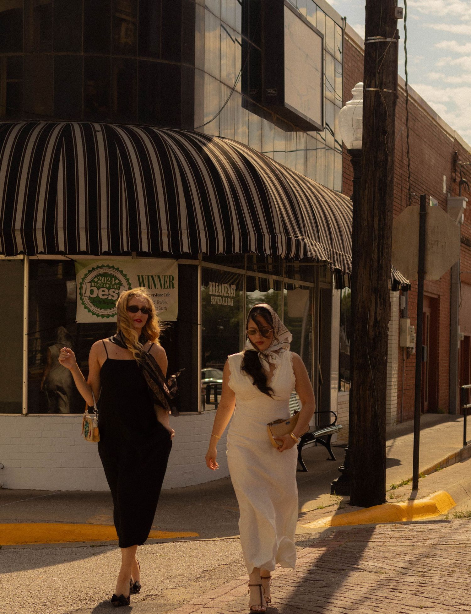 Two stylish women walking downtown sidewalk, one in black dress, one in white, past striped awning.