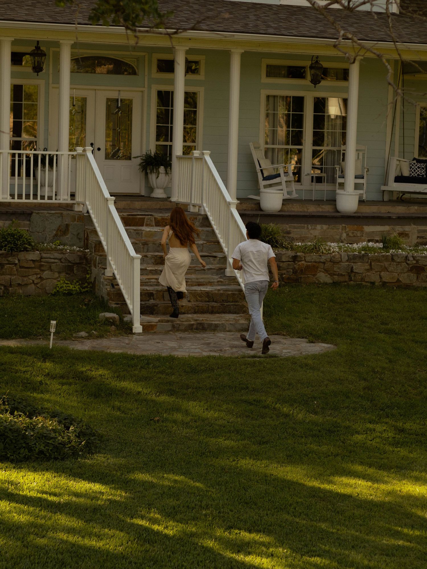 Two people walking up stone steps toward a charming green farmhouse with a wraparound porch on a sunny day.