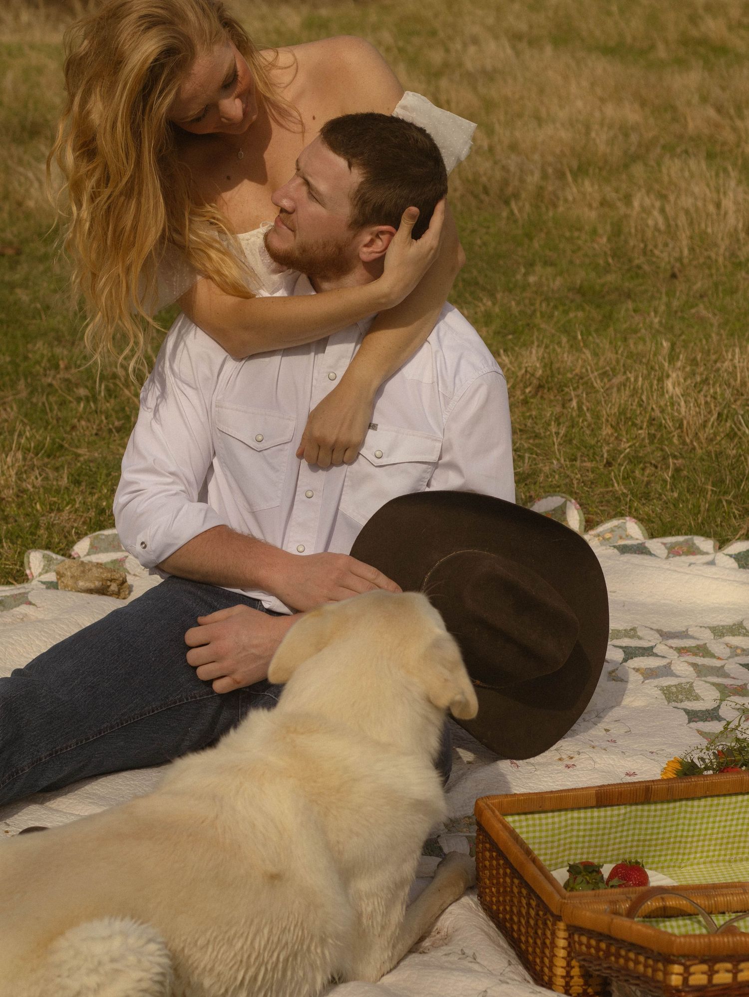 A couple shares a tender moment on a picnic blanket with their white dog in a sunny field.