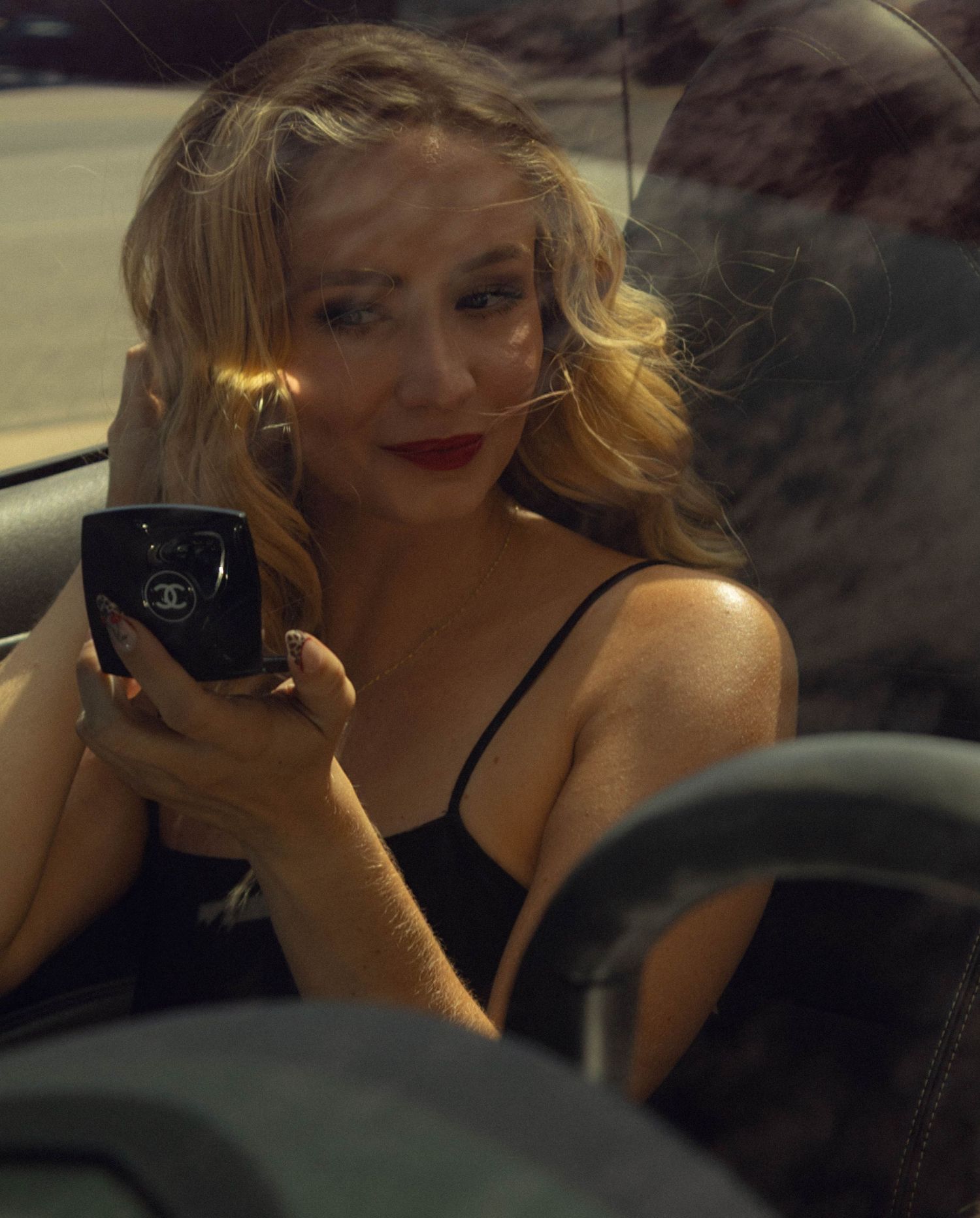 Blonde woman in black dress applying Chanel makeup in a convertible car, red lips, golden hour light.