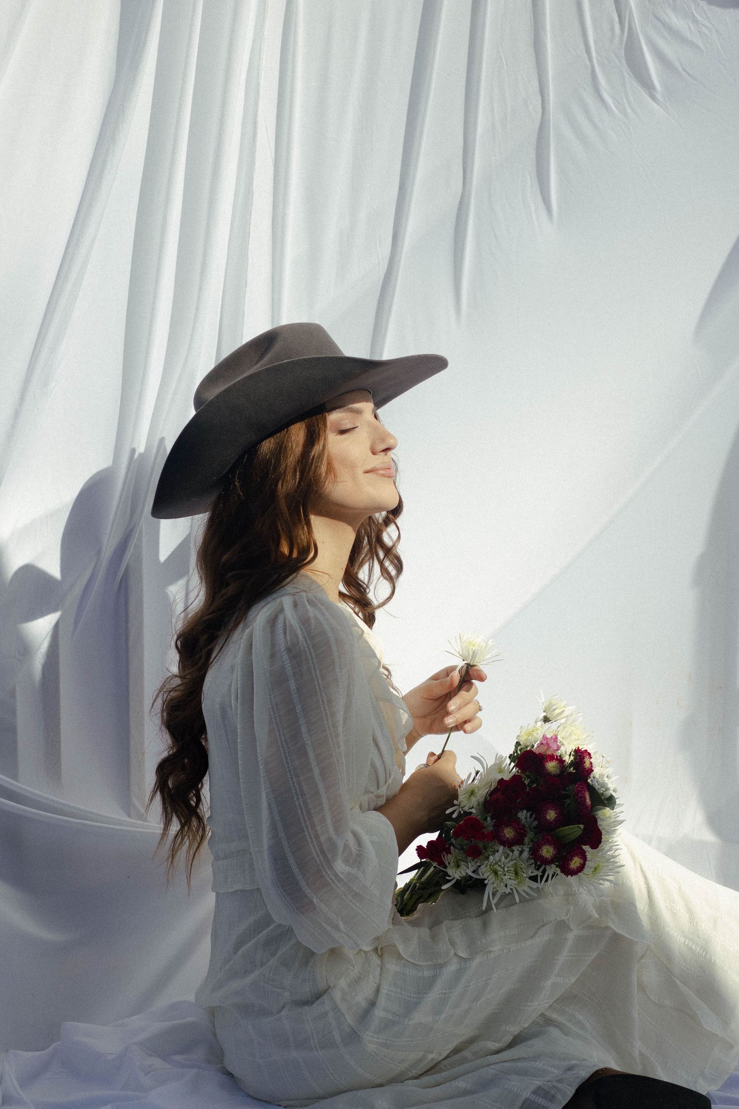 A person in a black hat and white dress poses with dark flowers against a light backdrop in a dreamy photo series.