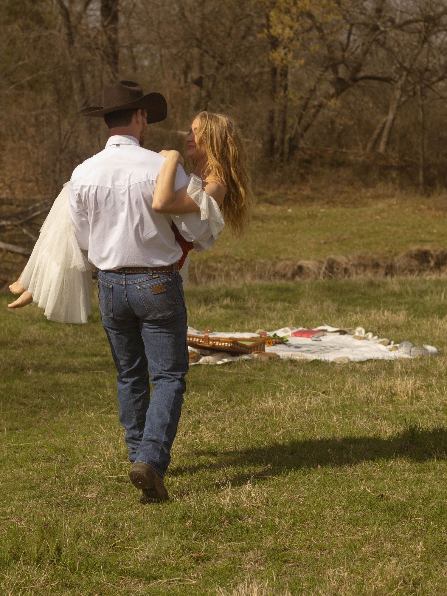 A cowboy in jeans and hat carries a woman in a white dress across a green field toward a picnic blanket.