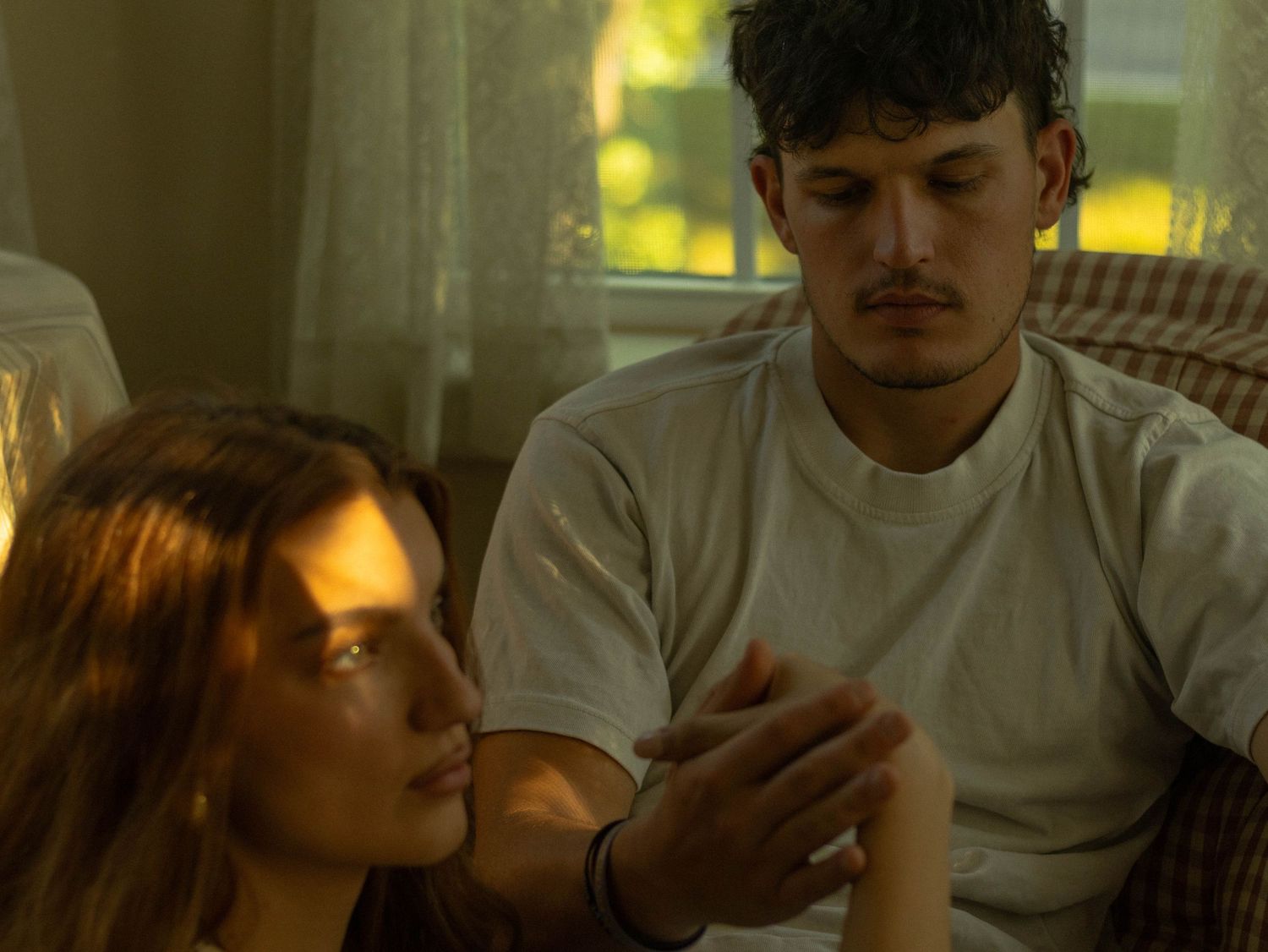 Young couple sitting together indoors in warm golden light, man gently touching woman's hand in intimate moment.