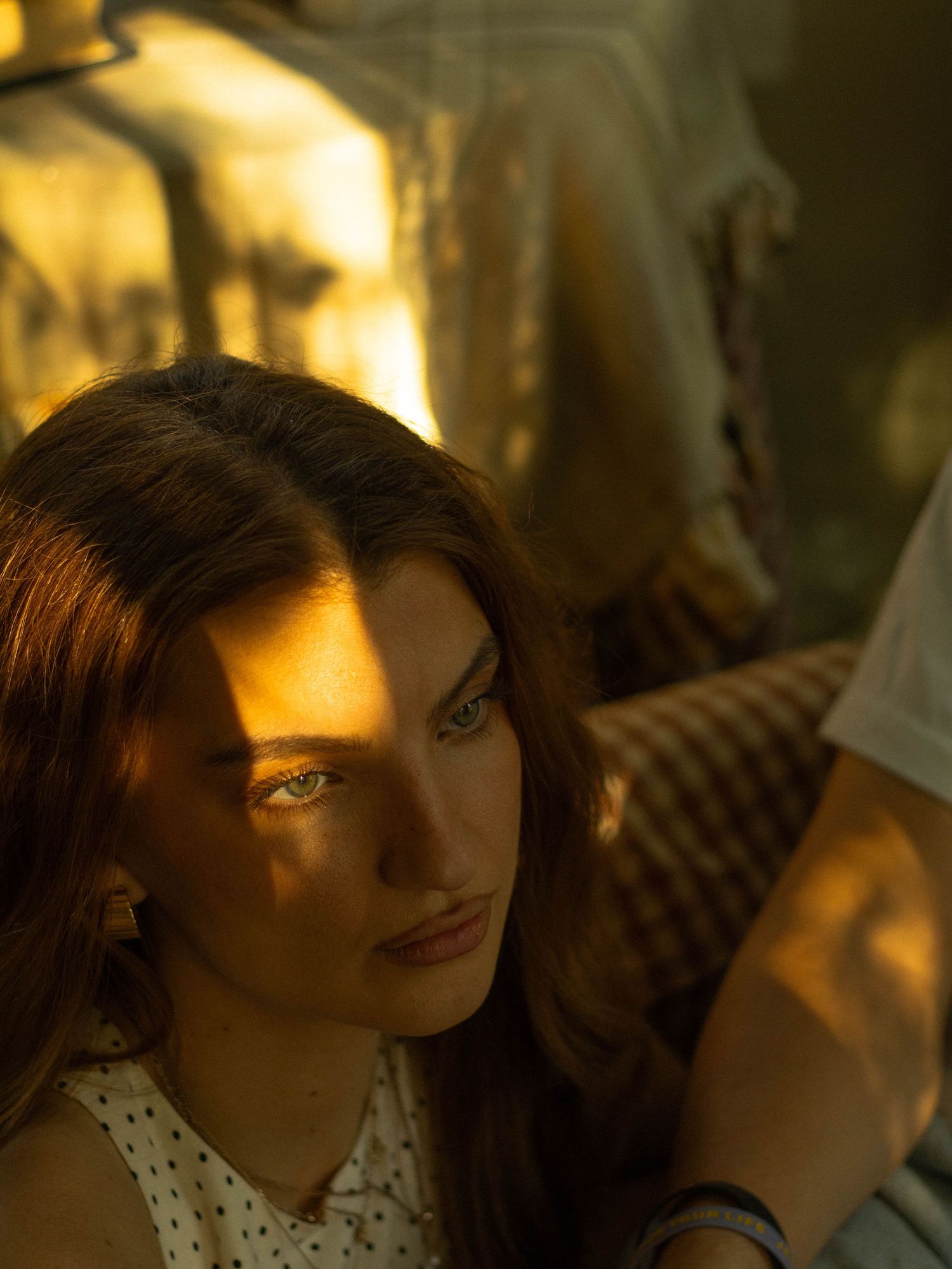 Young woman with auburn hair bathed in warm golden light, gazing thoughtfully in a moody outdoor setting.