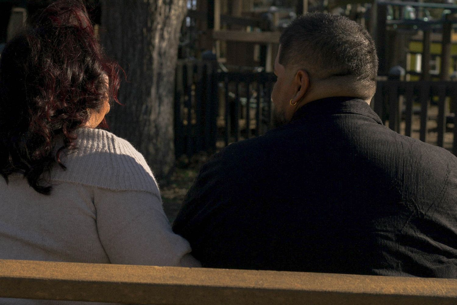 Two people's silhouettes blend with the evening shadows as they sit together on a park bench.