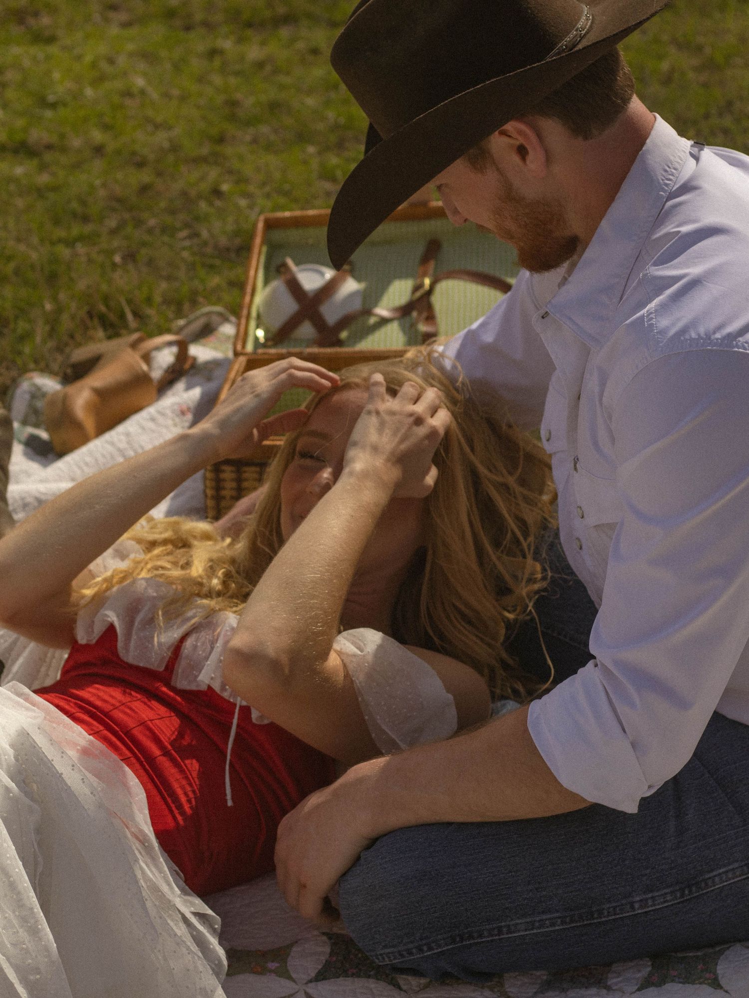 A couple in western attire sharing an intimate moment on the grass during a sunny day.