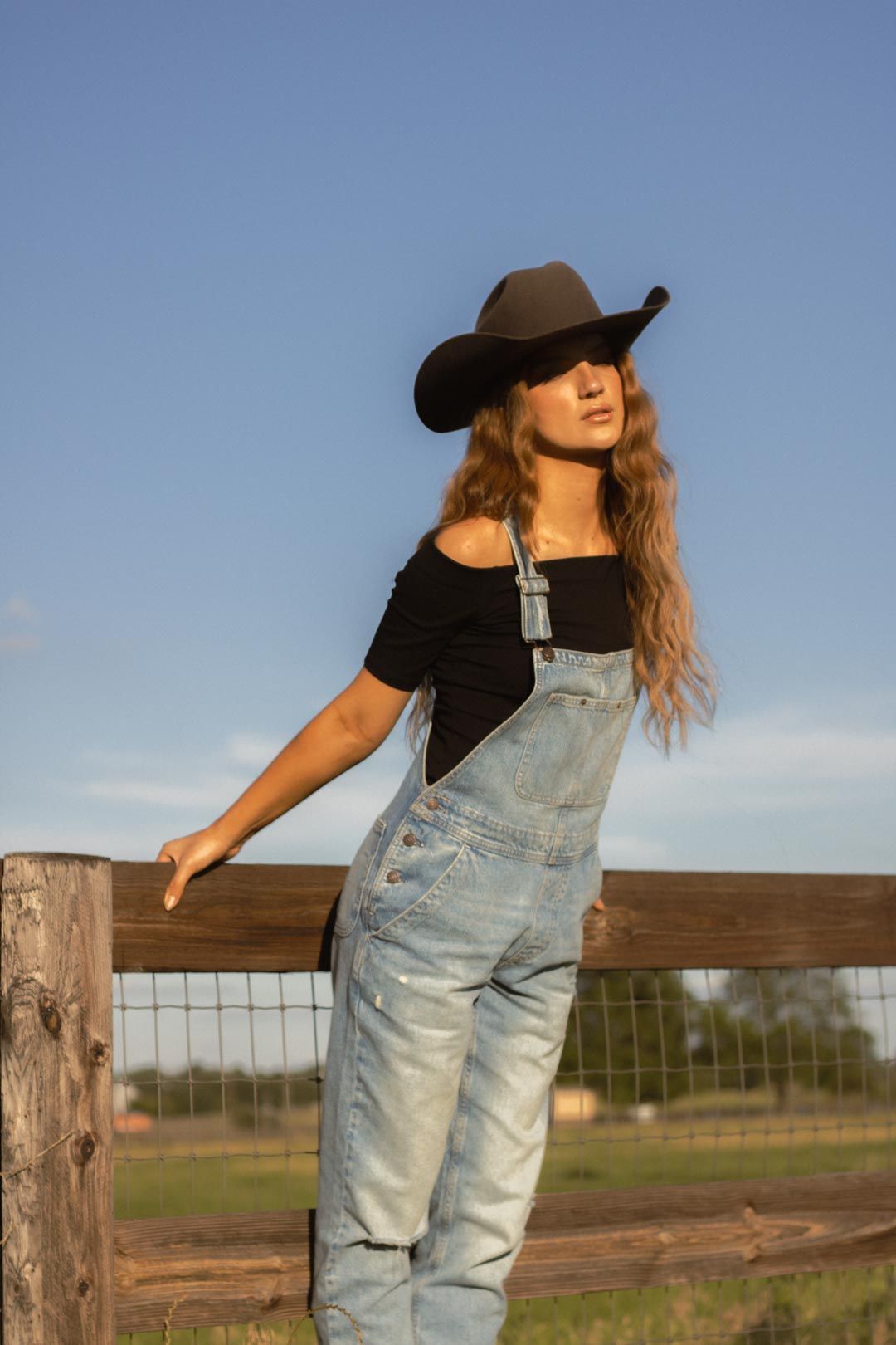 A figure in overalls leans against a wooden fence post in golden afternoon light.
