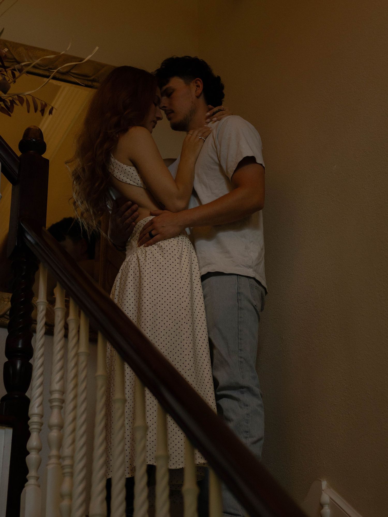 A couple shares a romantic kiss while embracing on a staircase railing in a warmly lit home.