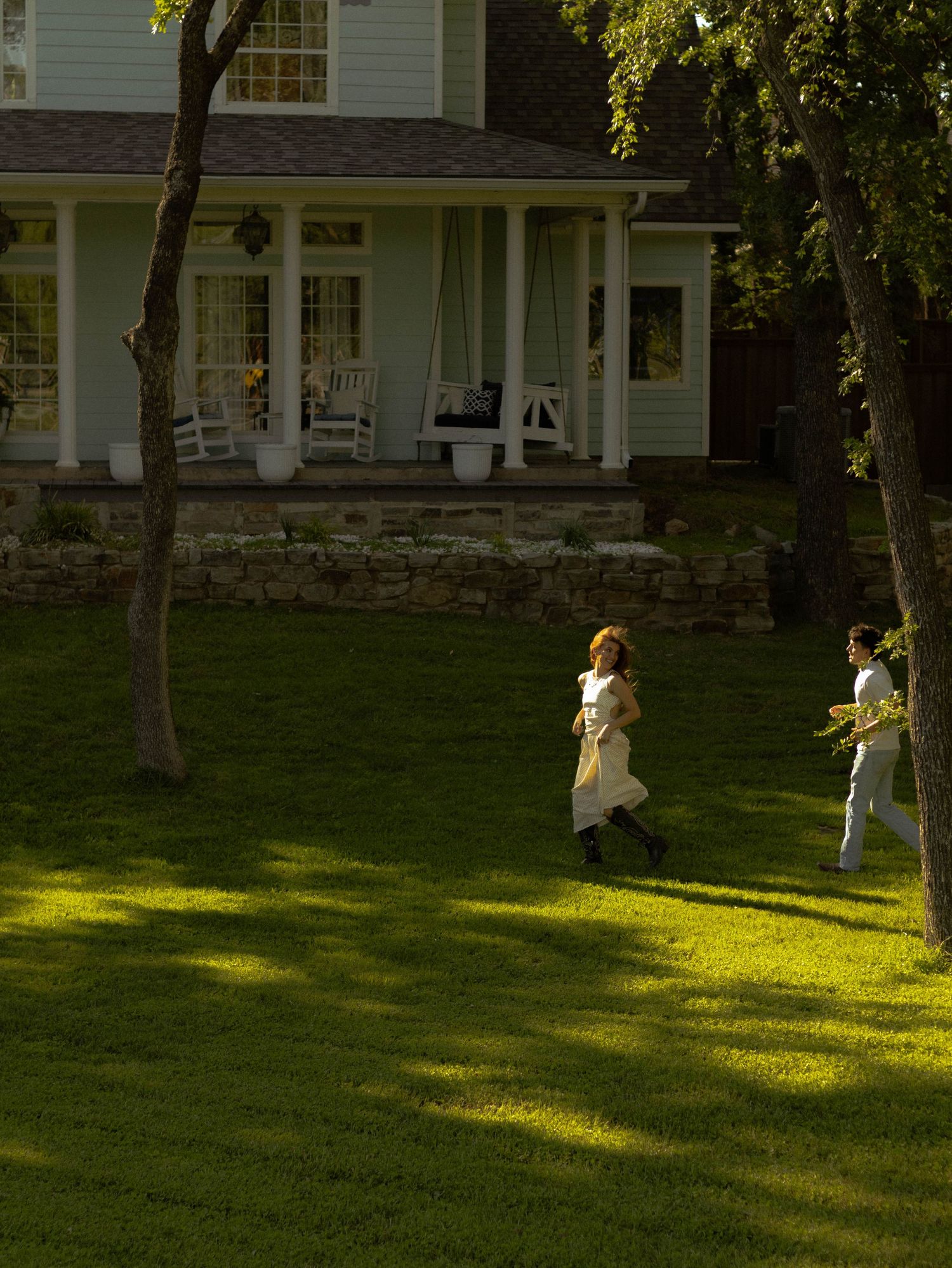 Two people running on lush green lawn in front of a charming white farmhouse with a wrap-around porch.