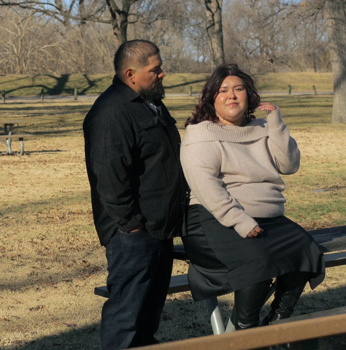 Two people sit together on a park bench surrounded by bare winter trees on a sunny afternoon.