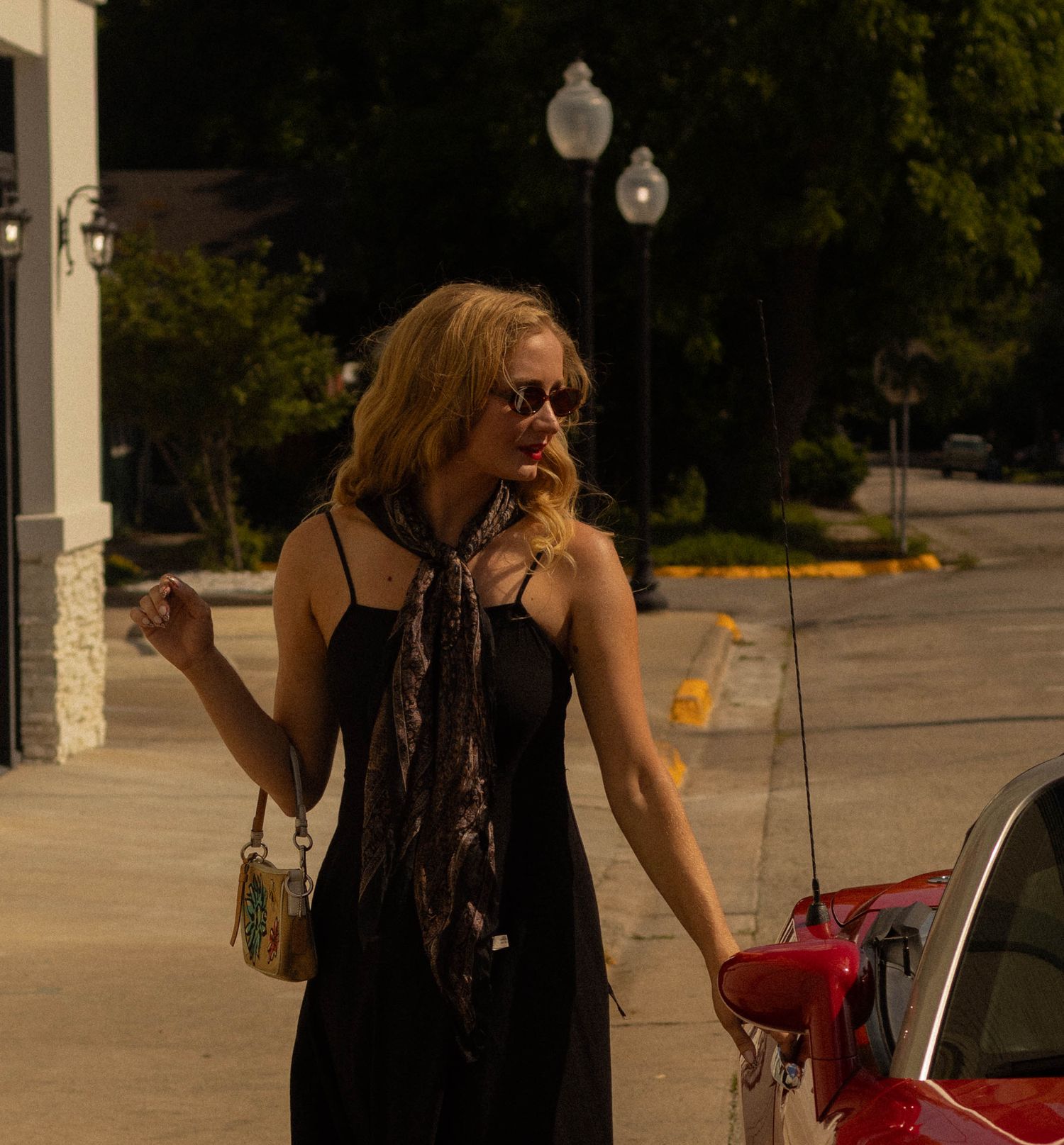 Blonde woman in black dress with leopard scarf standing beside a red convertible at a gas station.