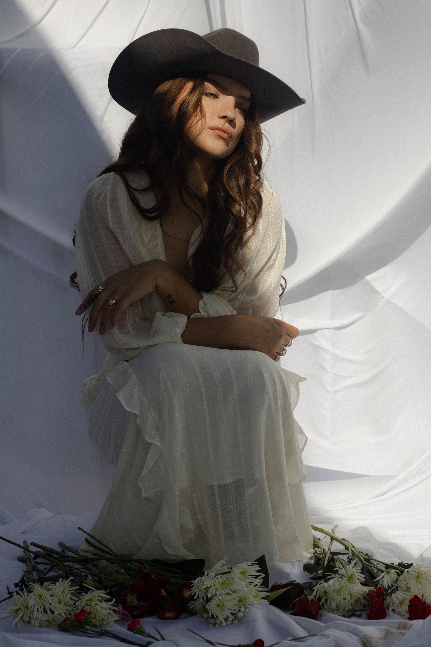 A person in a black cowboy hat and white dress poses against a white backdrop with scattered flowers on the ground.