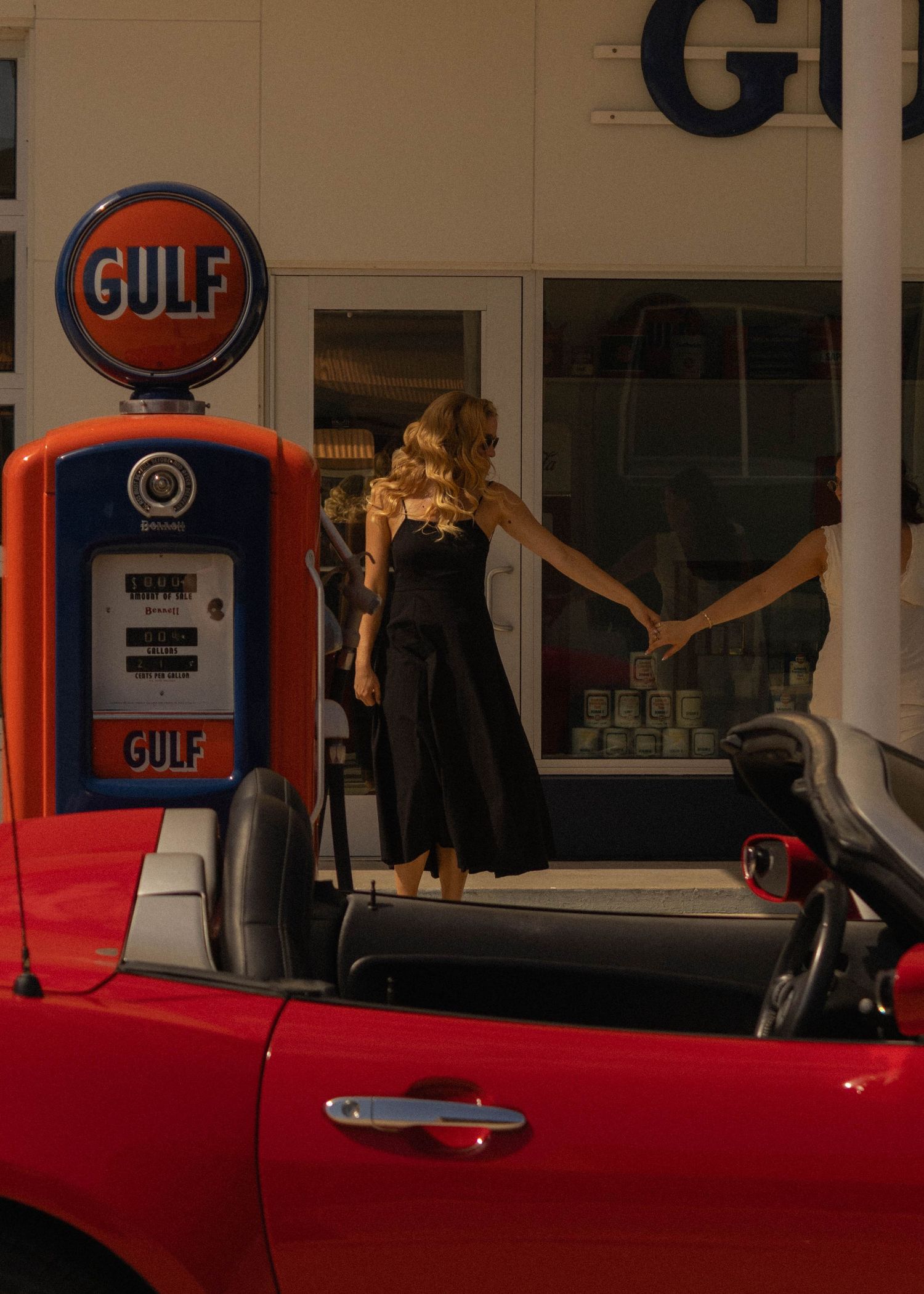 Woman in black dress standing in red convertible at vintage Gulf gas station with classic pump.