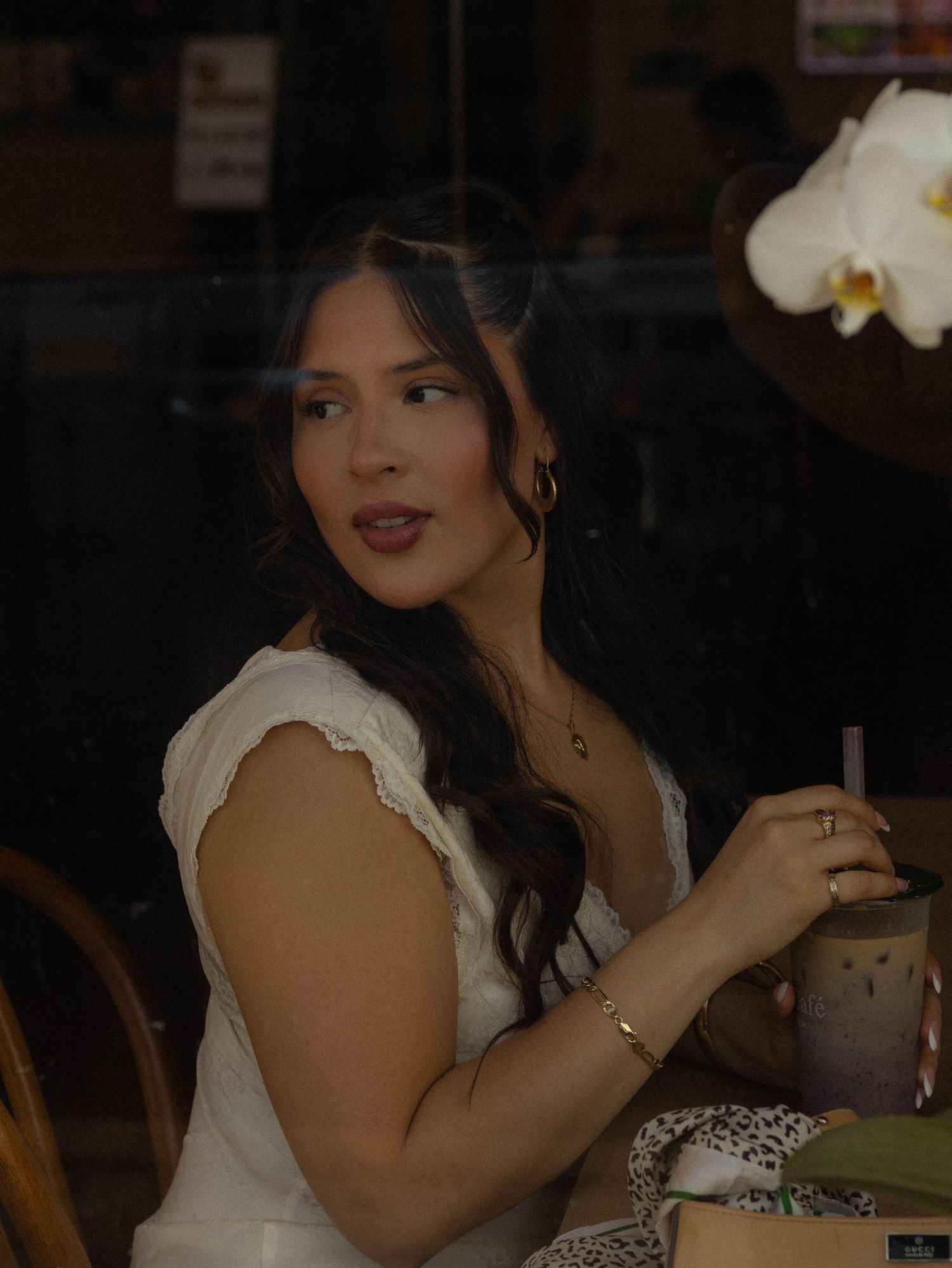 Young woman in white dress holding iced coffee drink, sitting at cafe table with white orchid flower nearby.