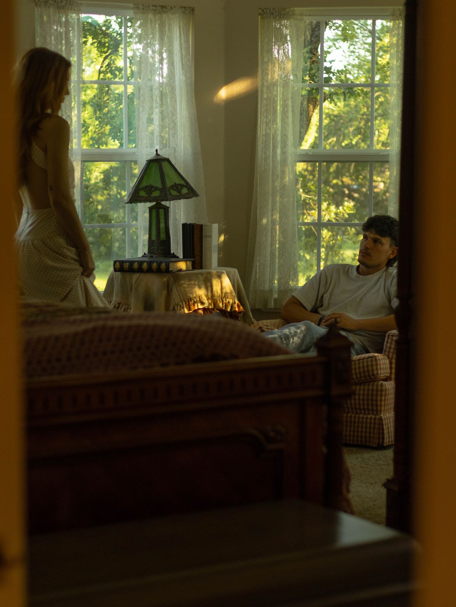 A man and woman in a warmly lit bedroom with lace curtains and a vintage Tiffany-style lamp.