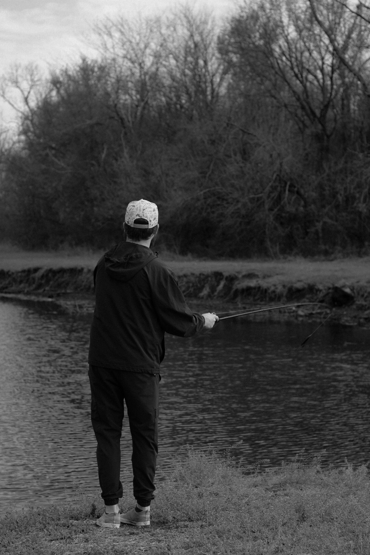 Person in dark clothing fishing by a lake on an overcast day.