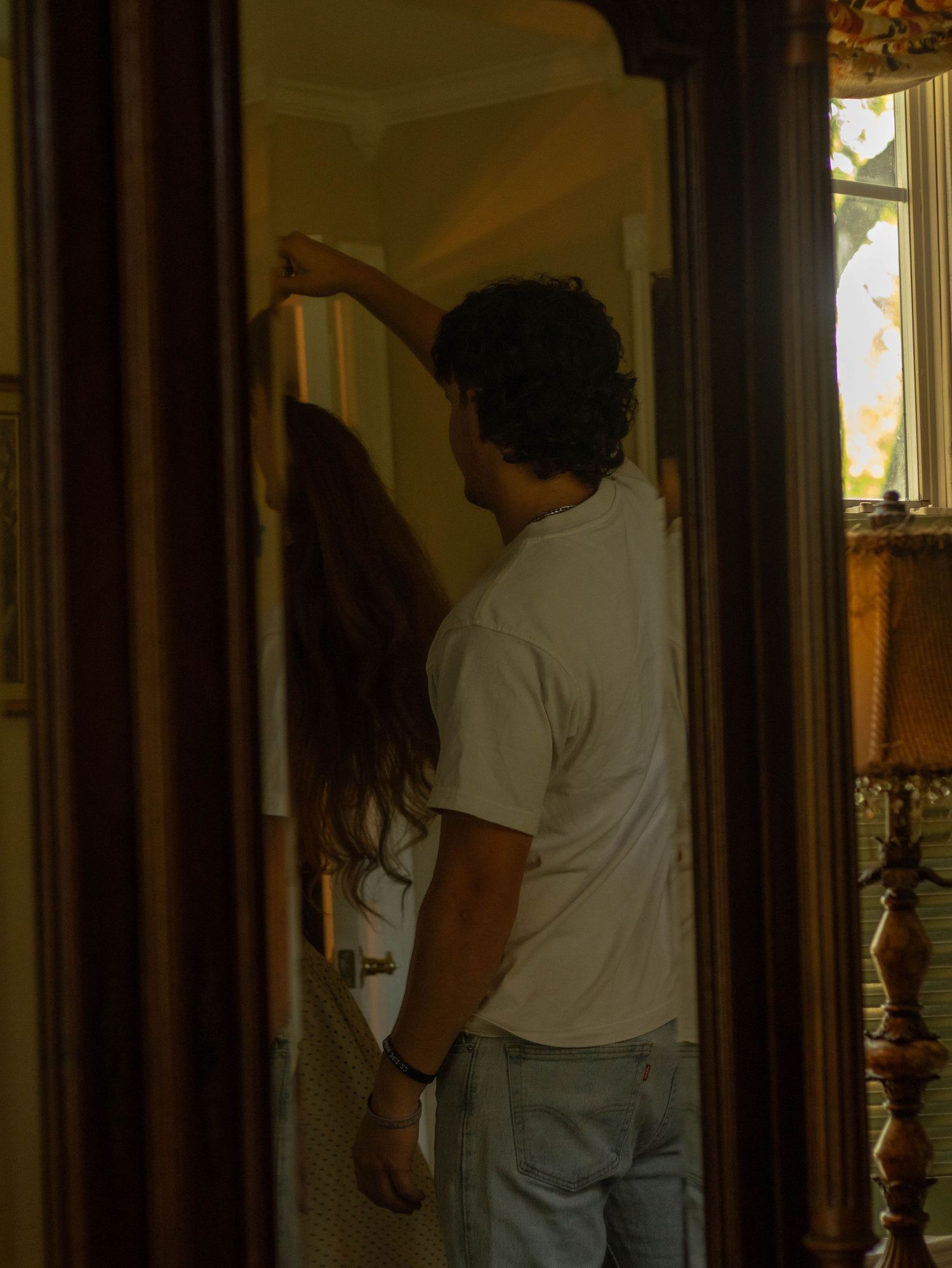 A couple stands together in a dimly lit doorway of an elegant home, the man in a white tee and jeans.