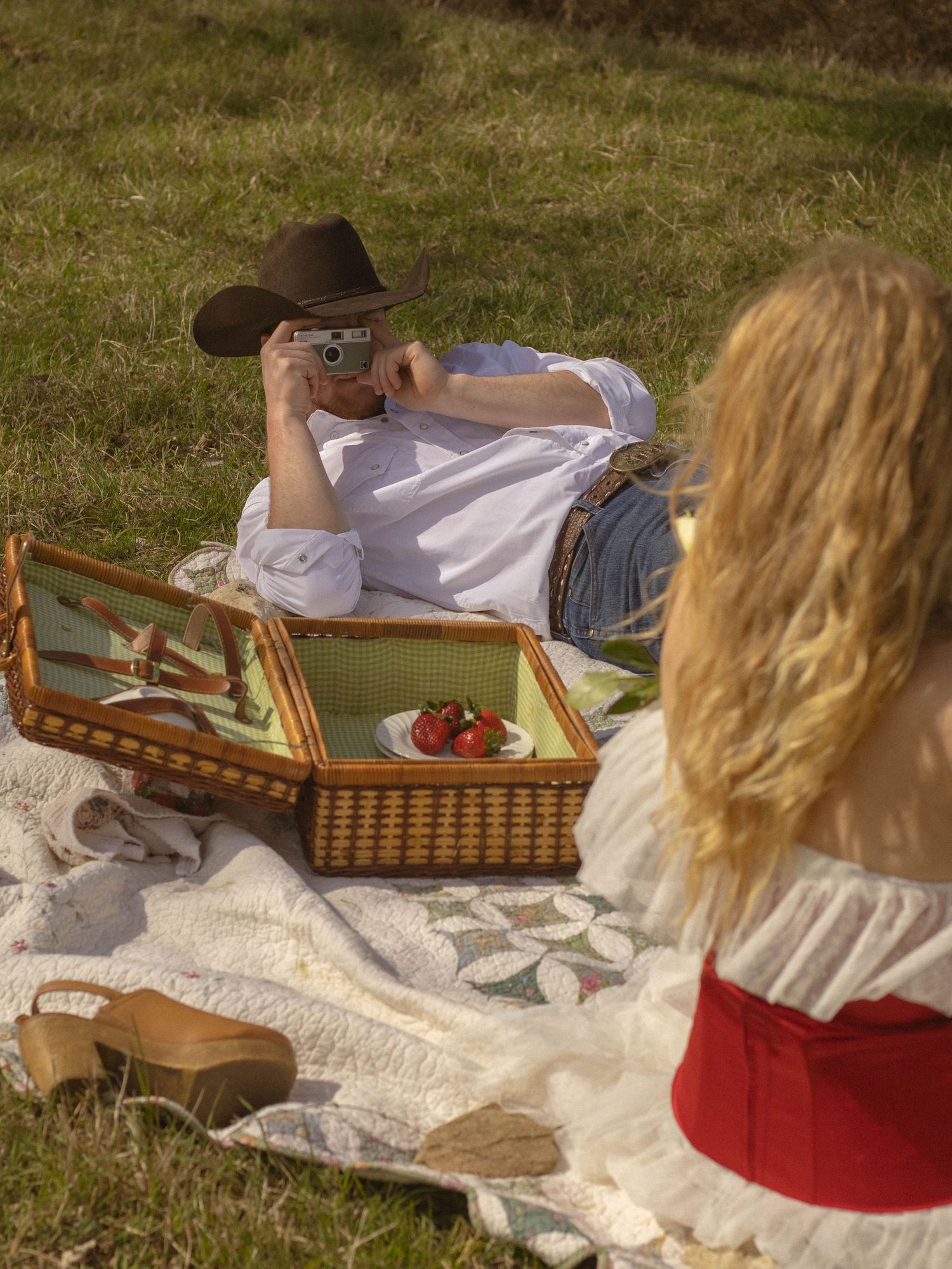 Couple enjoying a summer picnic with wicker basket and blanket on grassy meadow, capturing memories with camera.
