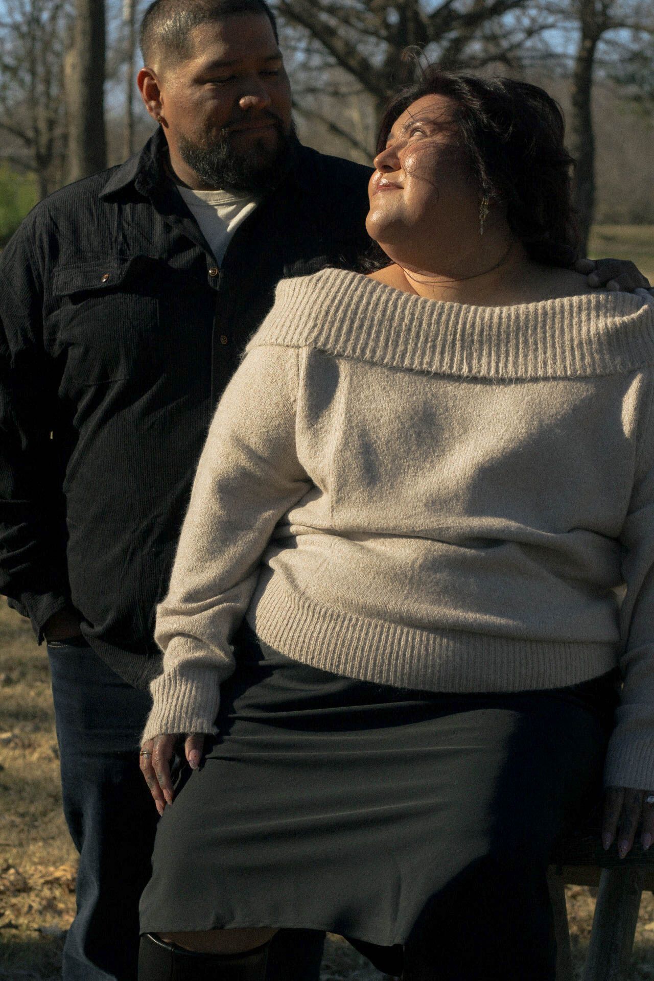 A couple shares an intimate moment outdoors with one wearing a cream sweater and the other in a black jacket.