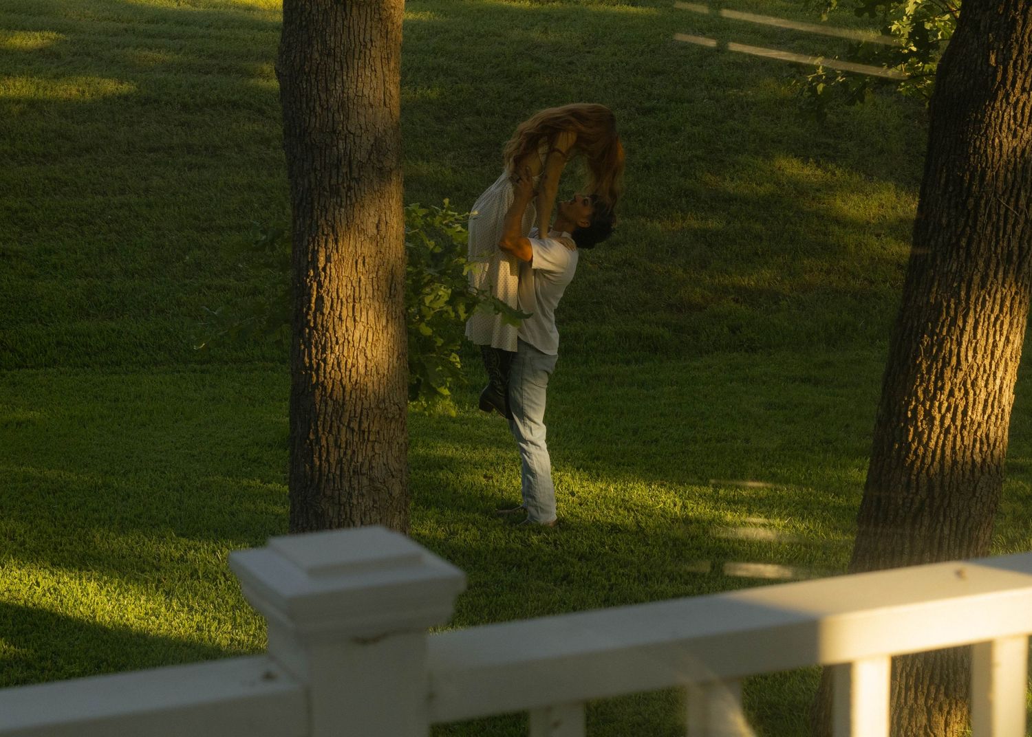 Red-haired woman walking between trees in sunlit green yard at dusk.