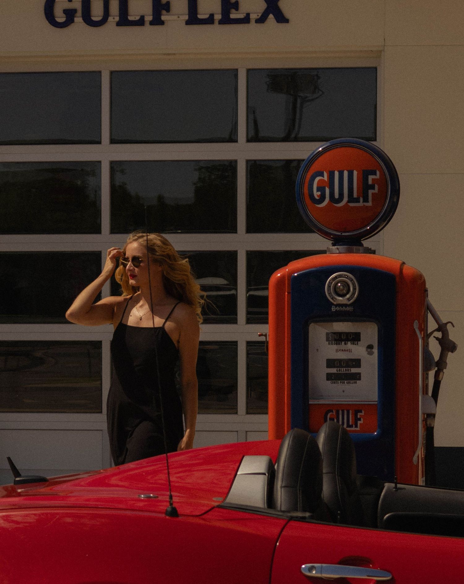 Woman in black dress poses by vintage Gulf gas pump at Gulflex station beside a red classic convertible car.