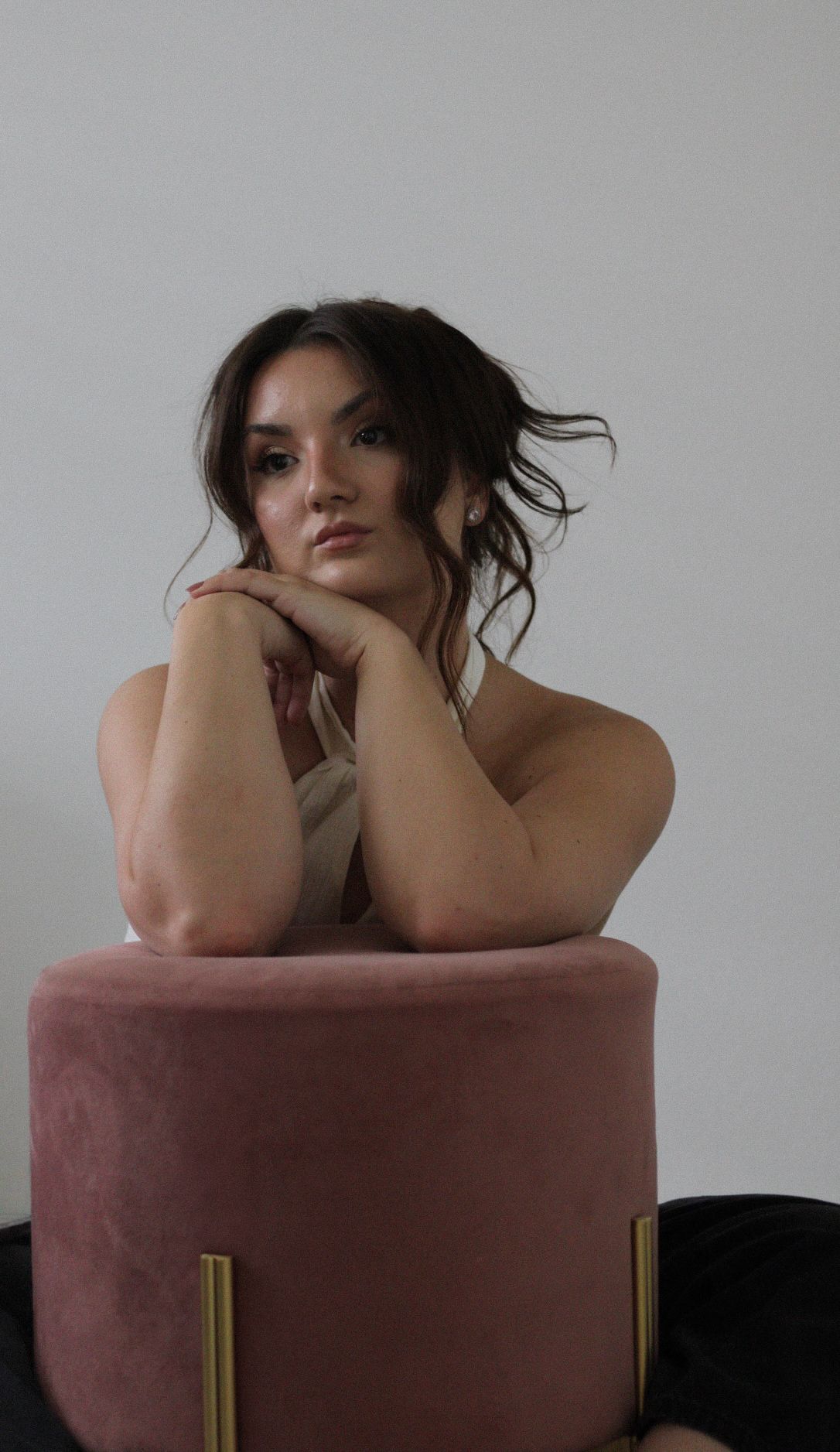 Portrait of someone posing on a pink velvet ottoman against a grey background.