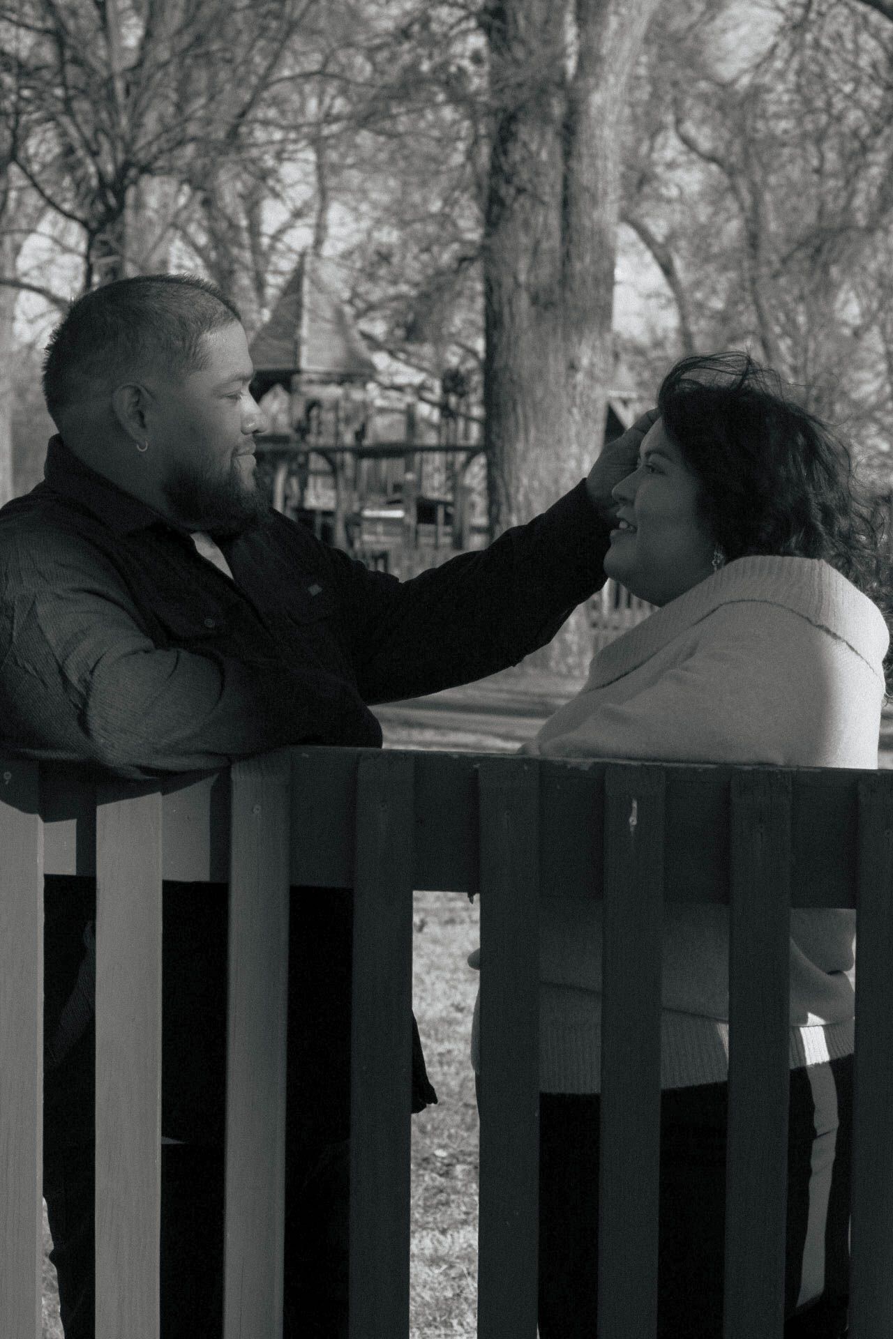 A couple interacts playfully behind wooden fence posts in a park setting.