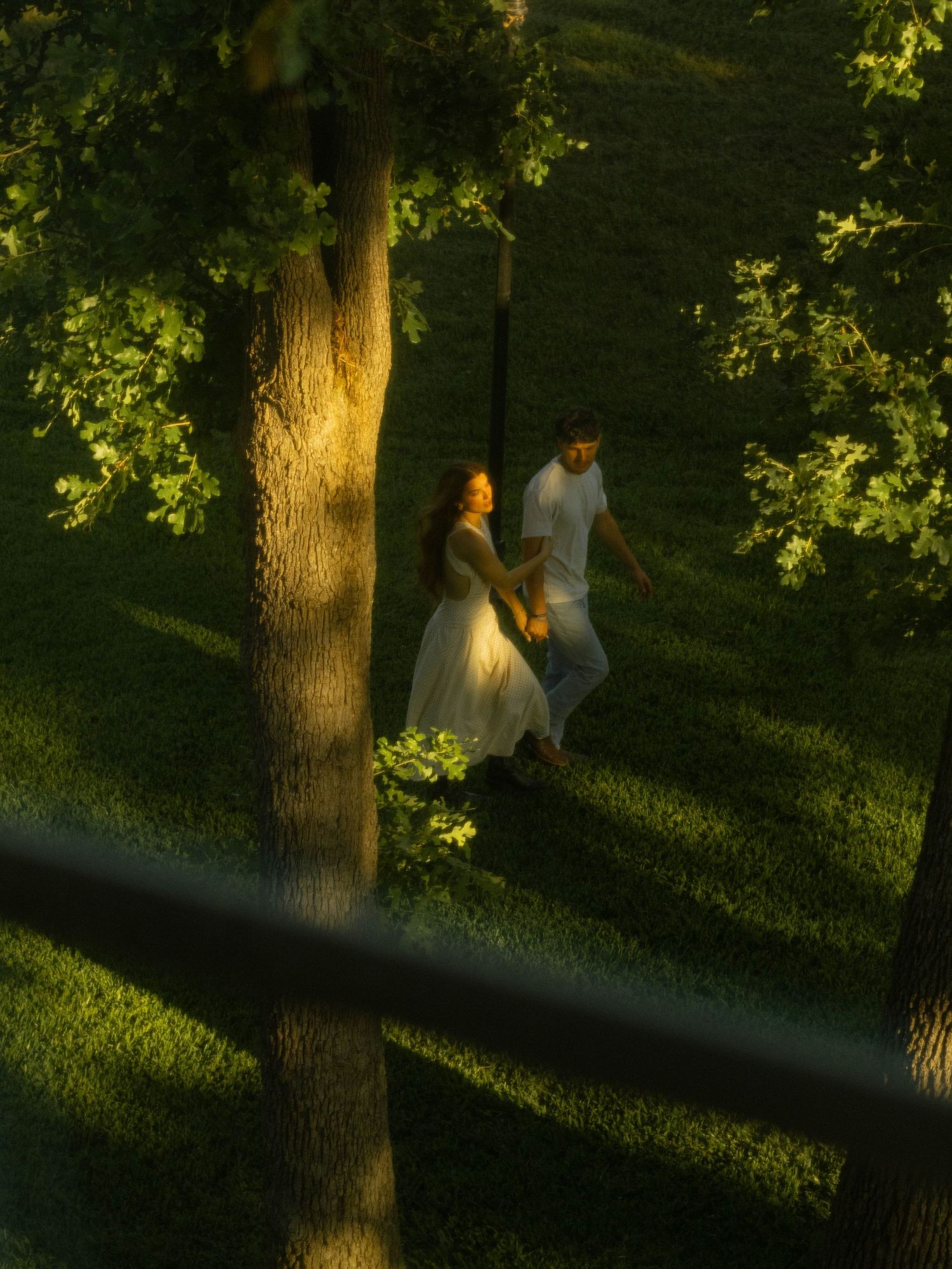A couple in white outfits walks hand in hand through a sunlit green park with tall trees casting shadows.