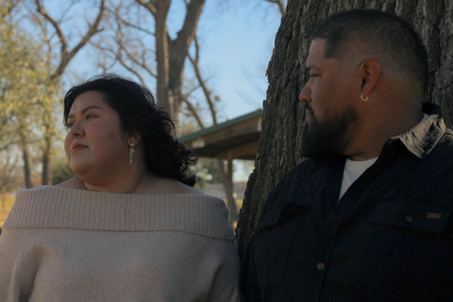A couple stands together against a tree trunk in a park during golden hour.