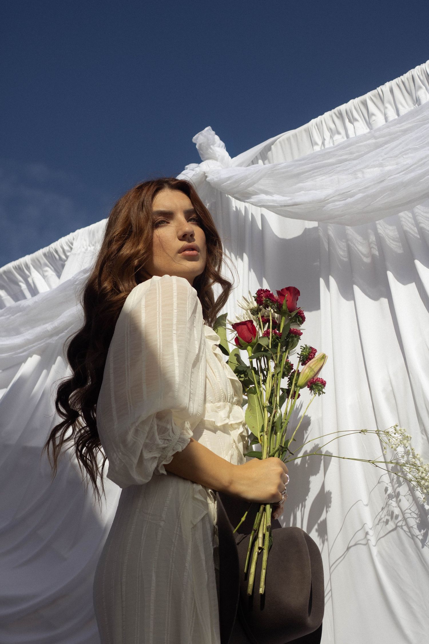 A person in a white dress holds red roses against a backdrop of billowing white sheets on a sunny day.