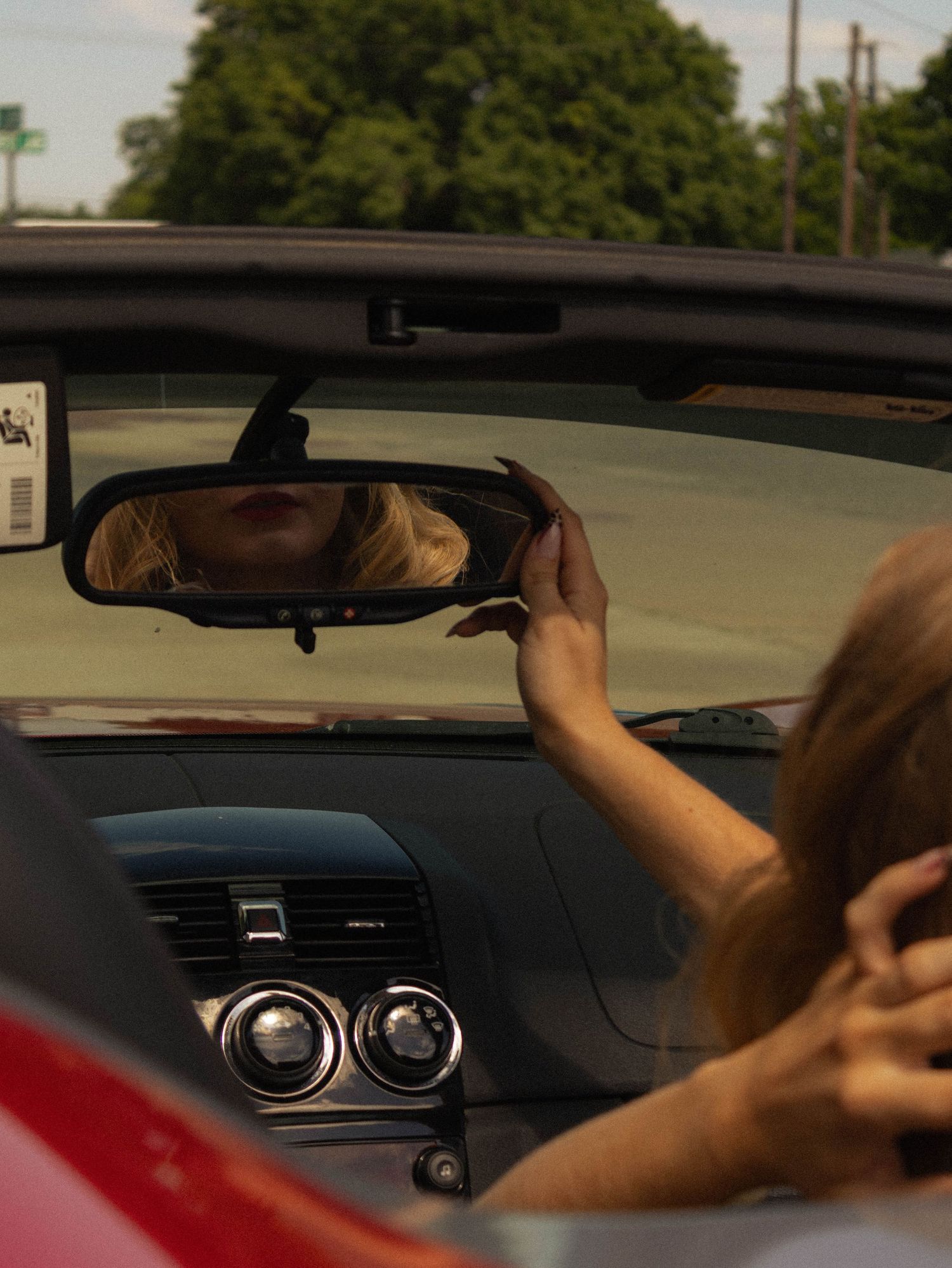 Blonde woman adjusting rearview mirror in red convertible car, reflection visible, sunny day outdoors.