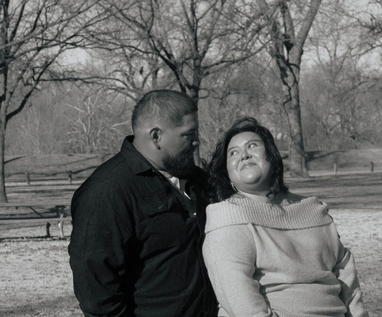 A couple embraces while sitting outdoors in a winter landscape with bare trees.