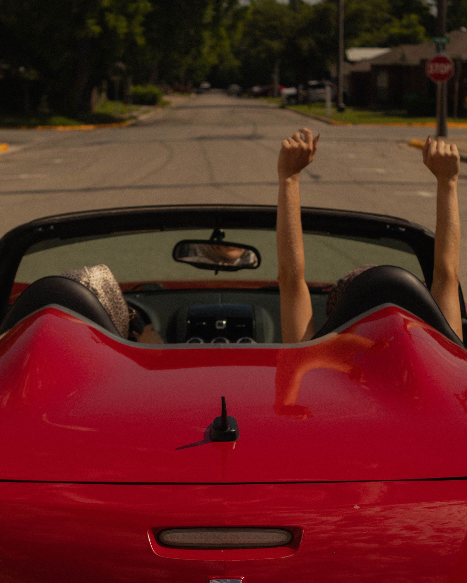 Two women celebrate with arms raised in a red convertible sports car on a sunny suburban street.
