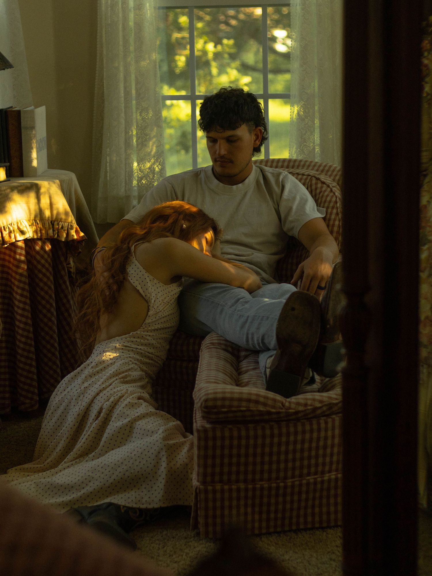 A couple relaxes in a plaid armchair by a sunlit window, woman in floral dress resting on man's lap.